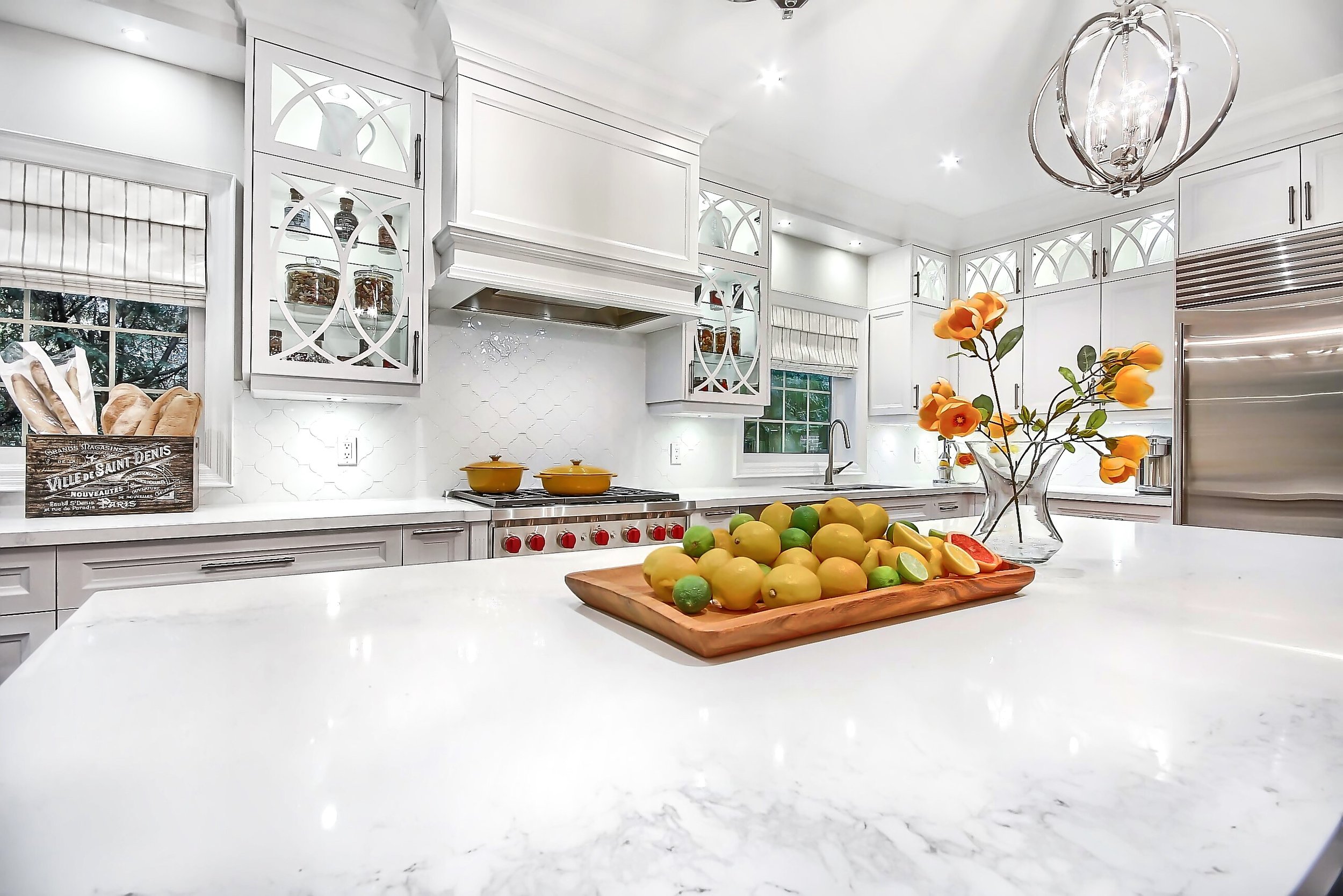 A white kitchen with a marble countertop, a bunch of lemons and limes in a wooden tray, a clear vase with orange flowers, white cabinets, a window with blinds, and a modern chandelier.