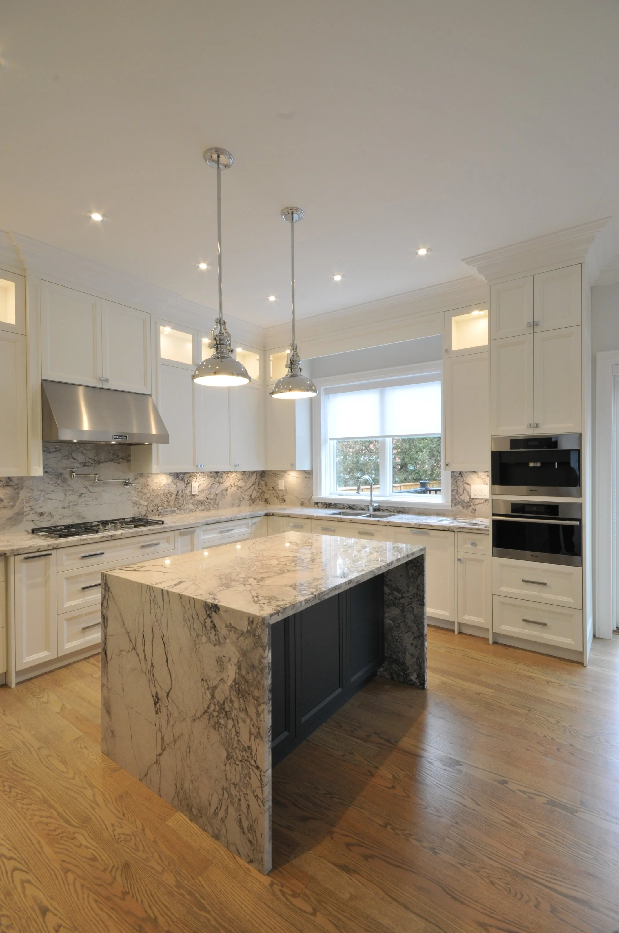 A modern kitchen with white cabinets, marble countertop, and a black island with matching marble surface. The kitchen has wood flooring, ceiling pendant lights, and a window above the sink.