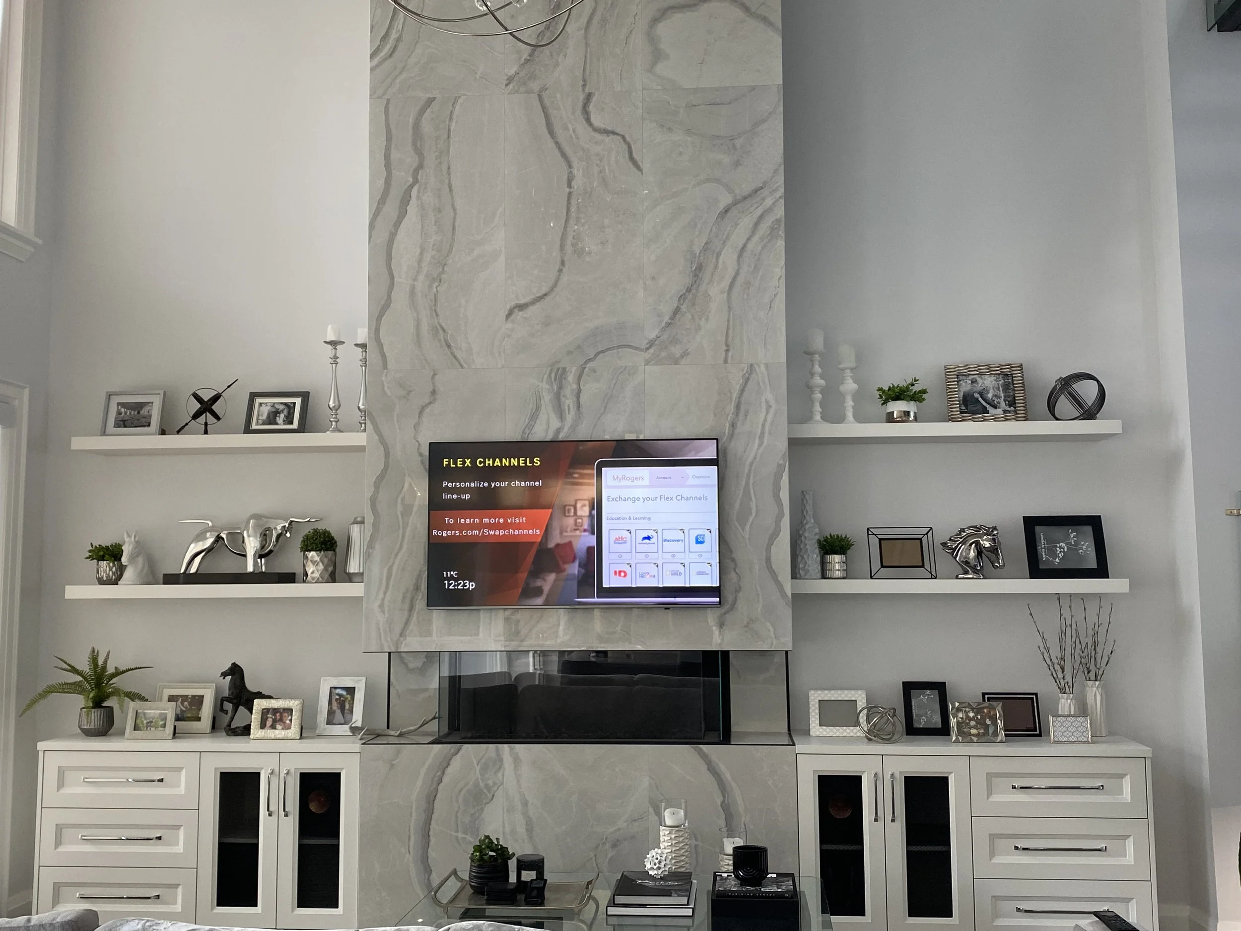 Living room with built-in white cabinets and shelves, decorated with picture frames, plants, decorative objects, and a mounted television on a marble wall.