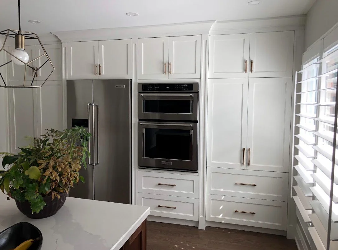 Modern kitchen with white cabinets, stainless steel refrigerator, double oven, and window with white blinds.