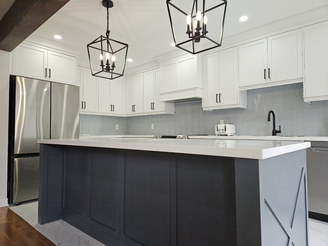 Modern kitchen with white upper cabinets, black hardware, a large gray island with a white countertop, stainless steel refrigerator, black faucet, white backsplash, and black hanging light fixtures.