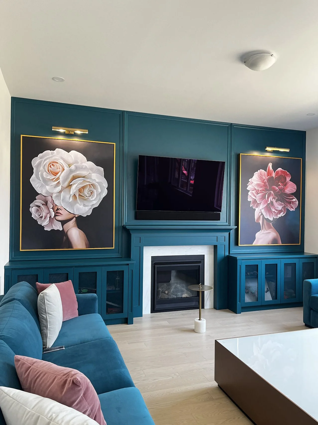Modern living room with teal built-in cabinets, two large floral portraits with backlit frames, a wall-mounted TV, a teal sofa with pink and white pillows, a white coffee table, and a small side table.