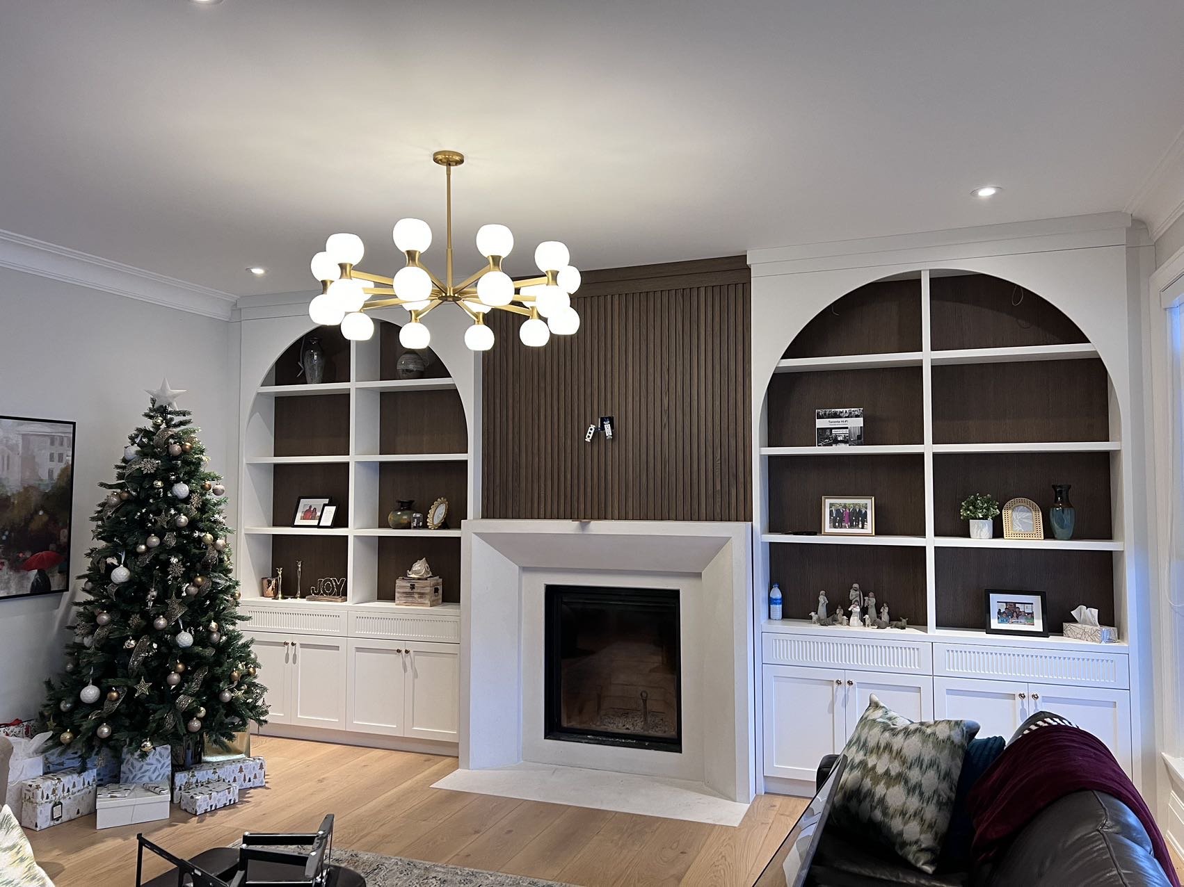 Living room decorated for Christmas with a Christmas tree, presents underneath, built-in shelves, a fireplace, a chandelier, and a couch with pillows.