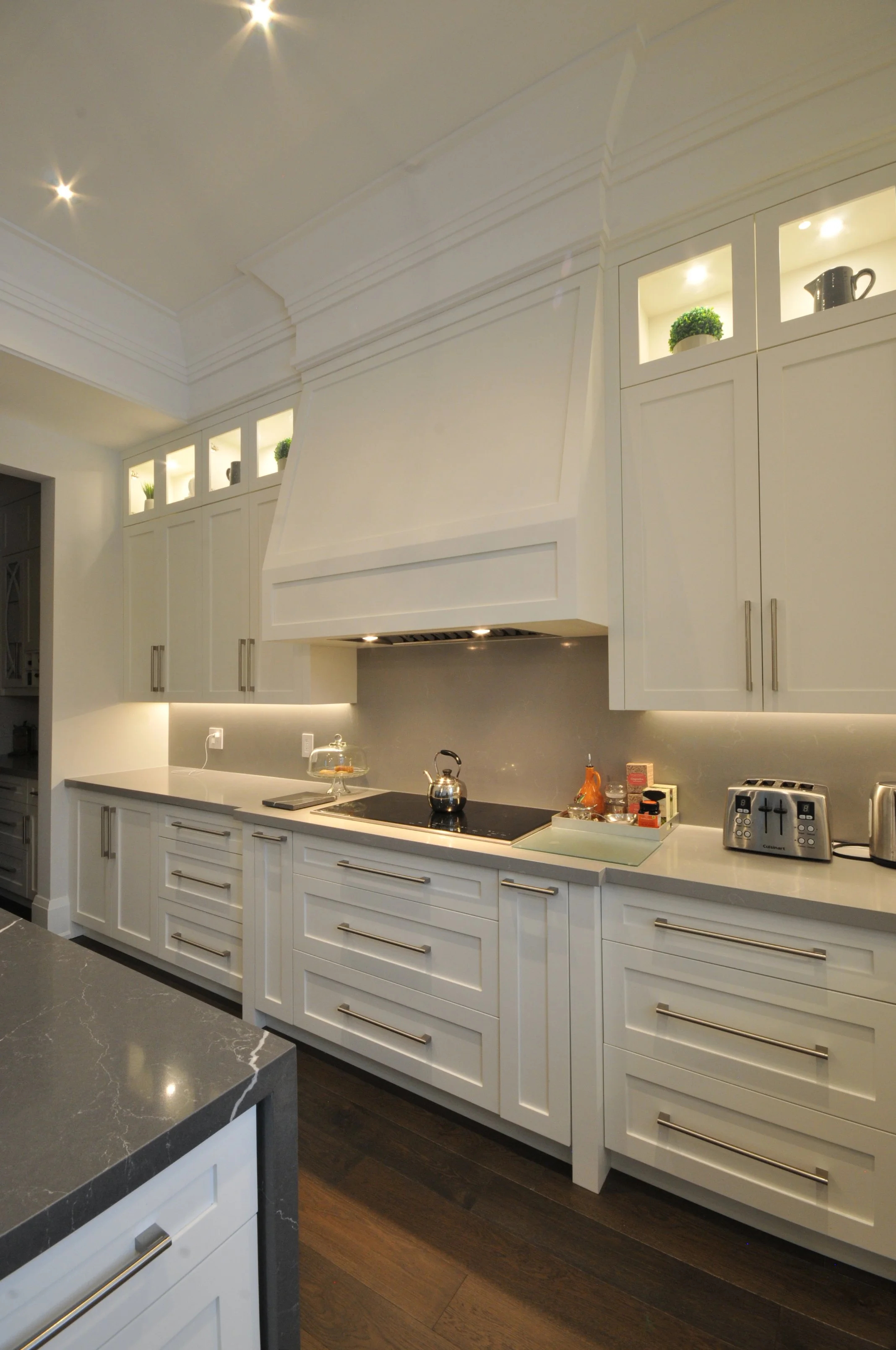 Modern kitchen with white cabinets, a gray countertop with a marble pattern, a black stovetop, and various small appliances and decor items on the counter, illuminated with built-in lighting.