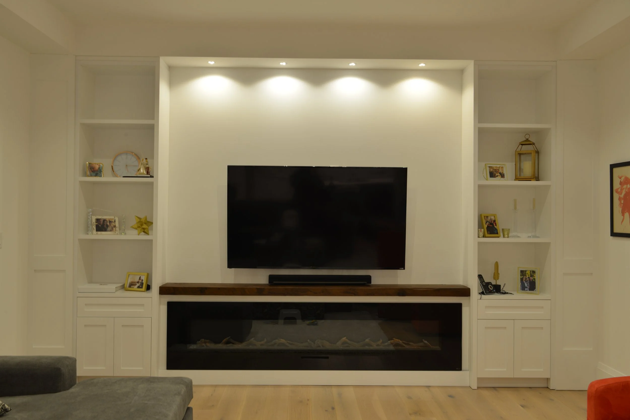 Living room entertainment wall with a large flat-screen TV, built-in white shelves with decorative items and framed photos, a fireplace underneath the TV, and hardwood flooring.