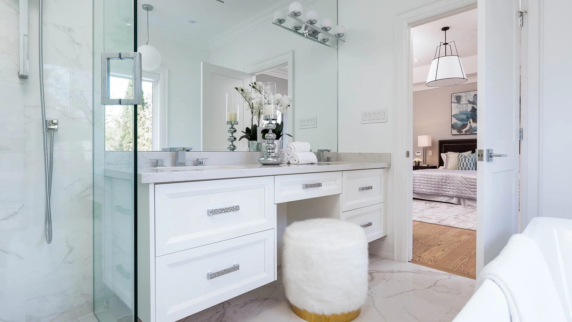 Modern white bathroom with marble floor, large mirror, and vanity with decorative hardware. Bathroom is bright with natural light, and there is a view into a bedroom with a bed, dressers, and artwork.