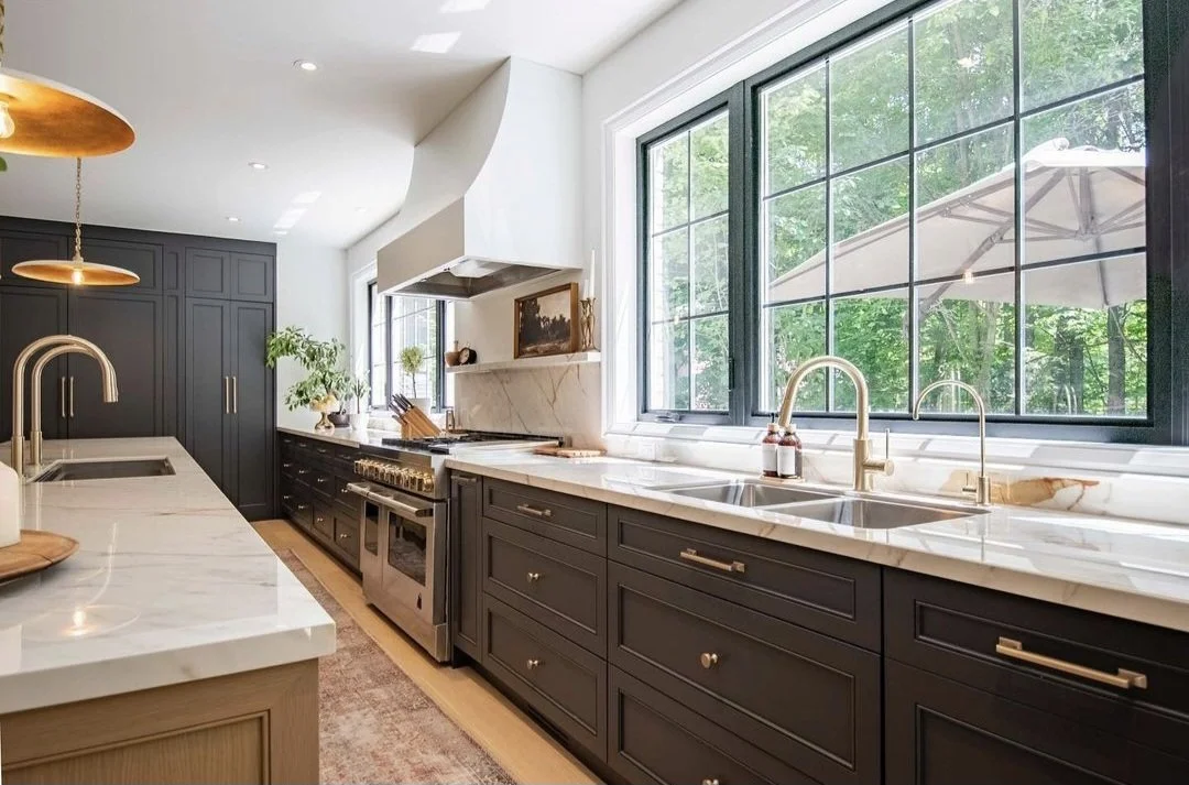 Modern kitchen with black cabinetry, white marble countertops, large window with a view of green trees, under-cabinet lighting, and a stainless steel stove with a gold range hood.