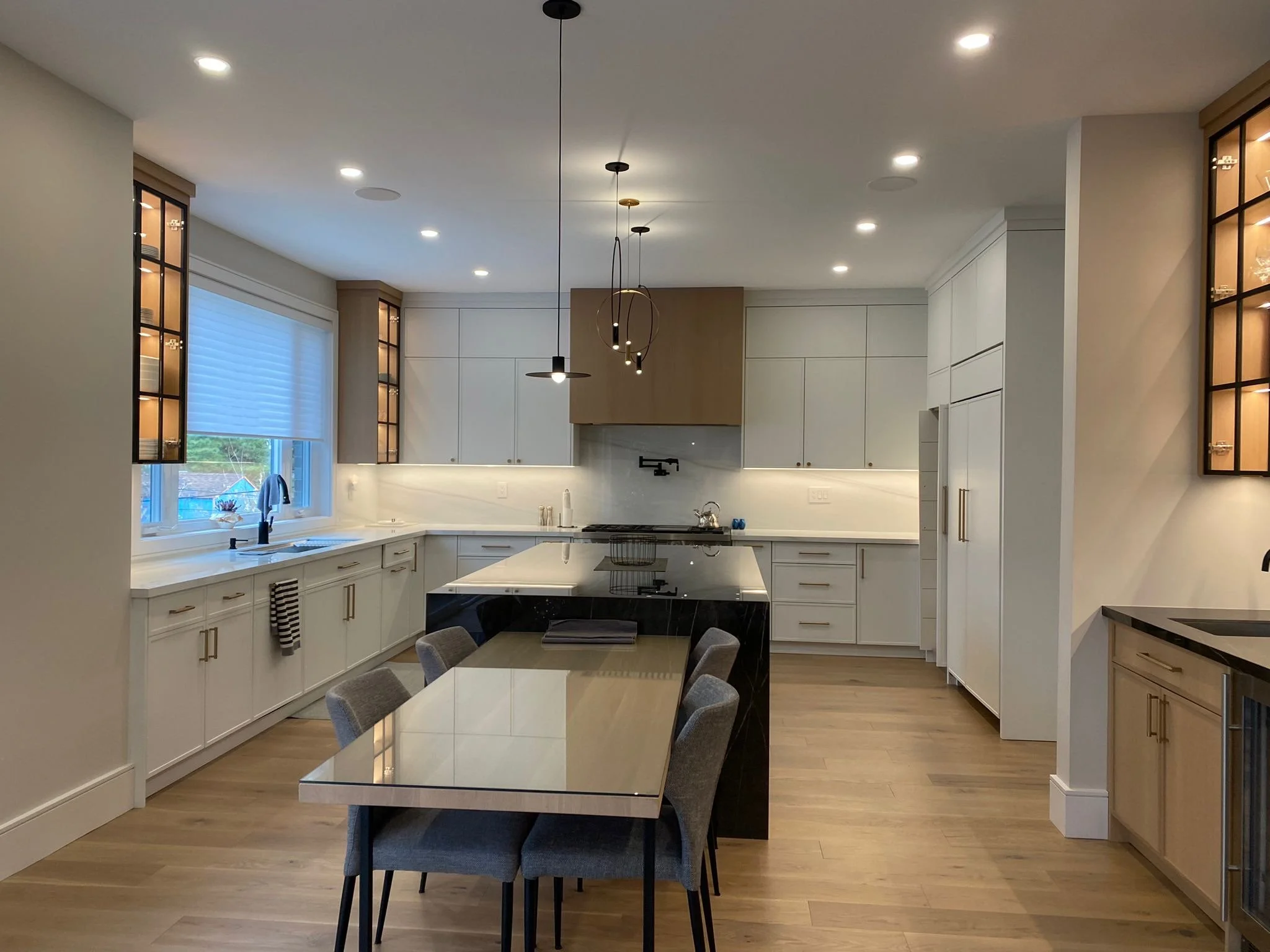 Modern kitchen with white cabinets, black hardware, and a large dining table with gray upholstered chairs. Features pendant lighting and a window with blue shades.
