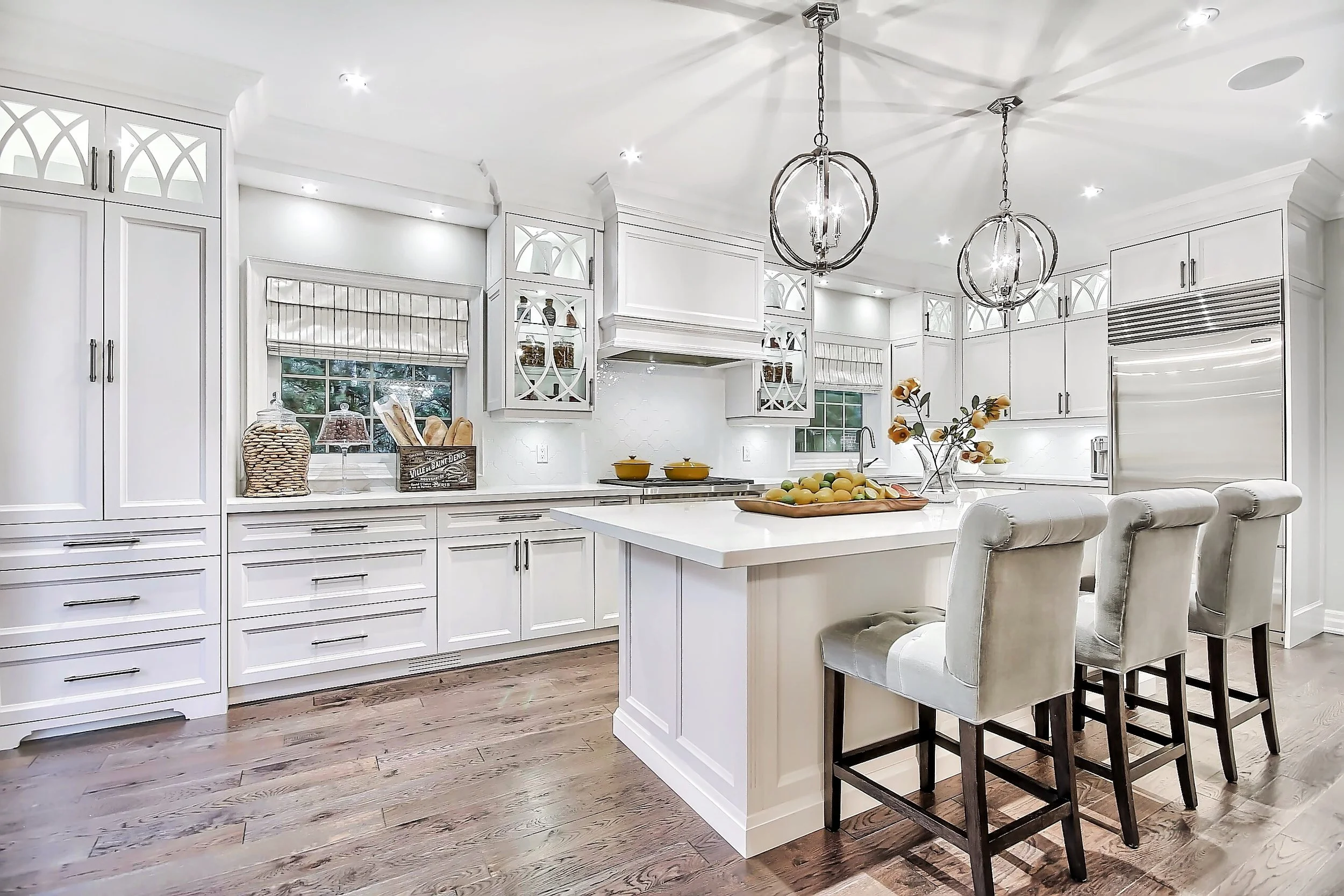 Modern white kitchen with island, pendant lights, and bar stools, featuring wood floors and decorated with fruits and jars.