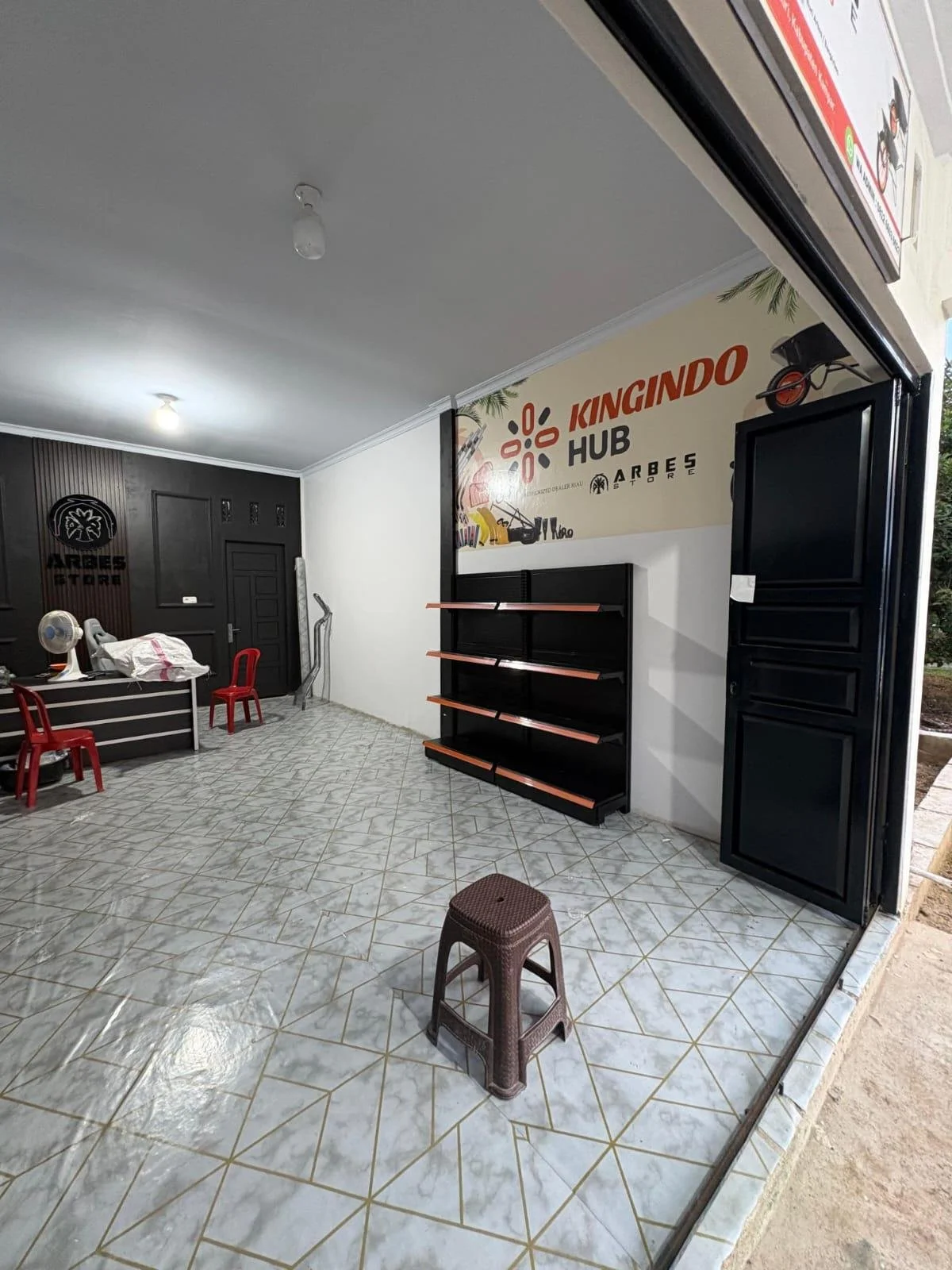 Empty storefront with tiled floor, black shelves, and a sign that reads 'KINGINDO HUB' with smaller texts and images related to Arbes Store.