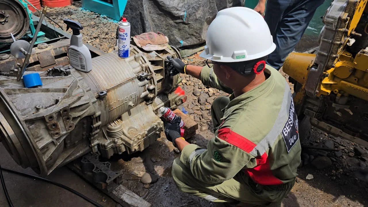 A worker in a safety uniform and hard hat inspecting a large industrial machine on a rocky surface, with tools and cleaning supplies nearby.