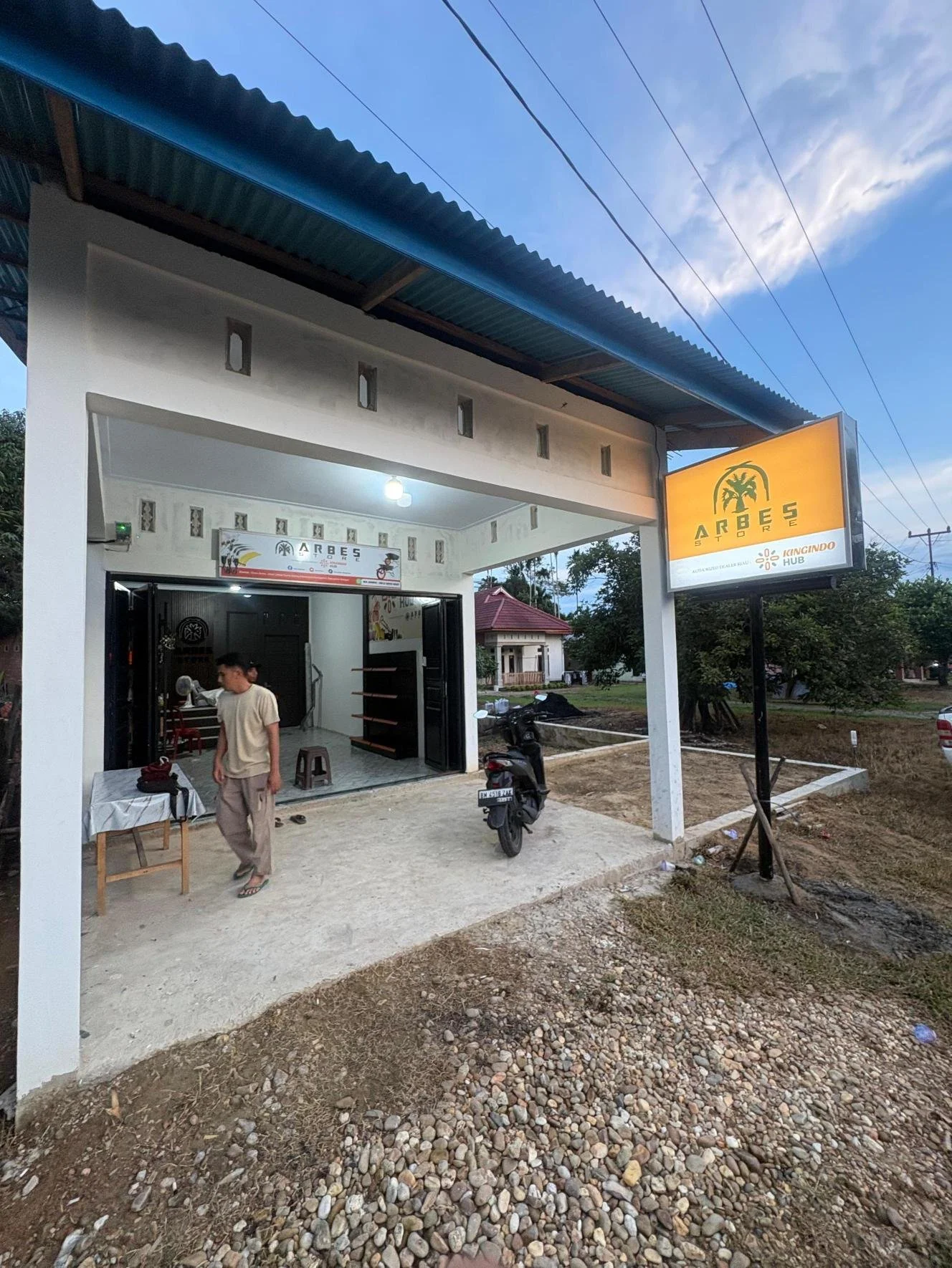 Exterior of a small store named 'Arbes Store' with a yellow sign, a motorcycle parked outside, and a person walking near a table inside the store, during daytime.