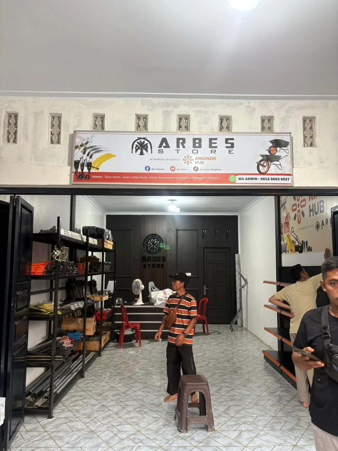 Inside a store with shelves of various items and people browsing. A man in a striped shirt stands near a small stool, while others look at products on the right side.