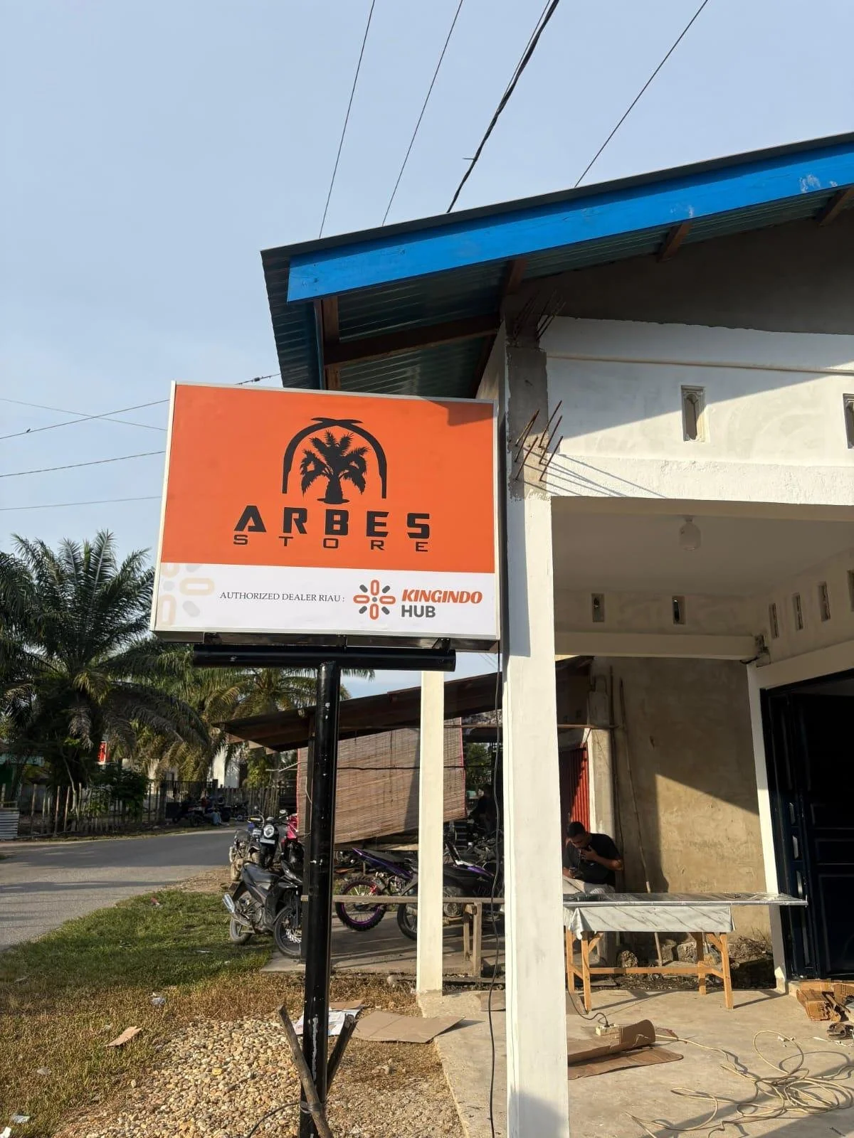 Sign for Arbes Store in front of a building, with motorcycles parked nearby, and a man sitting outside, in a sunny outdoor setting.