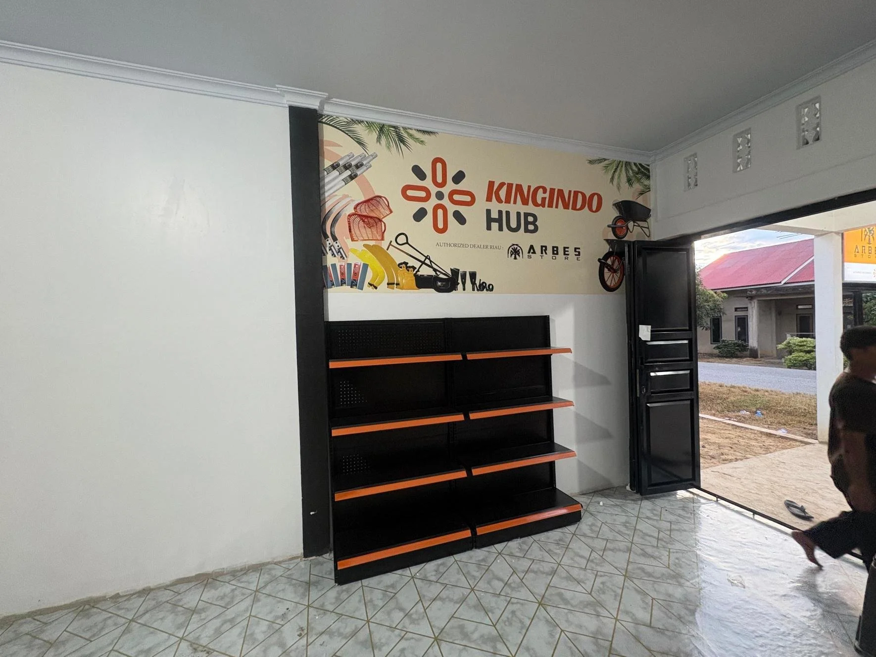 Interior of a store with a blank black and orange shelving unit, a wall sign that reads 'Kingindo Hub' with illustrations of tools and bikes, partially open door showing a street outside, and a person on the right side of the image.