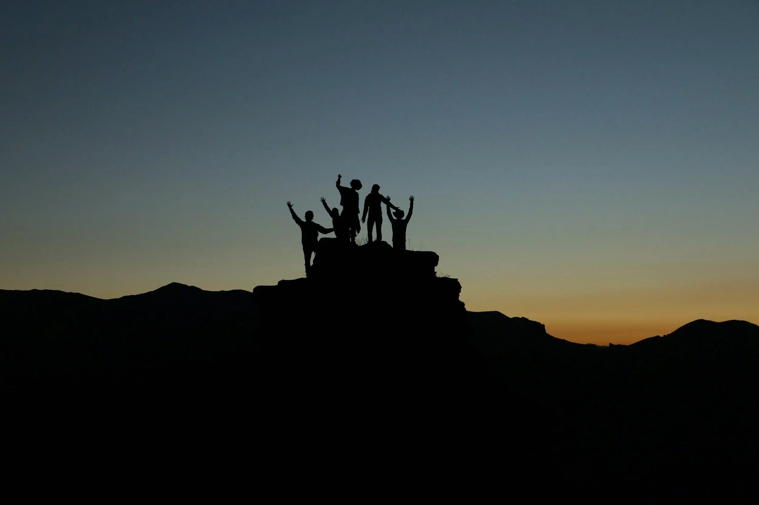 Silhouettes of five people standing on a rock formation at sunset, celebrating with their hands raised.