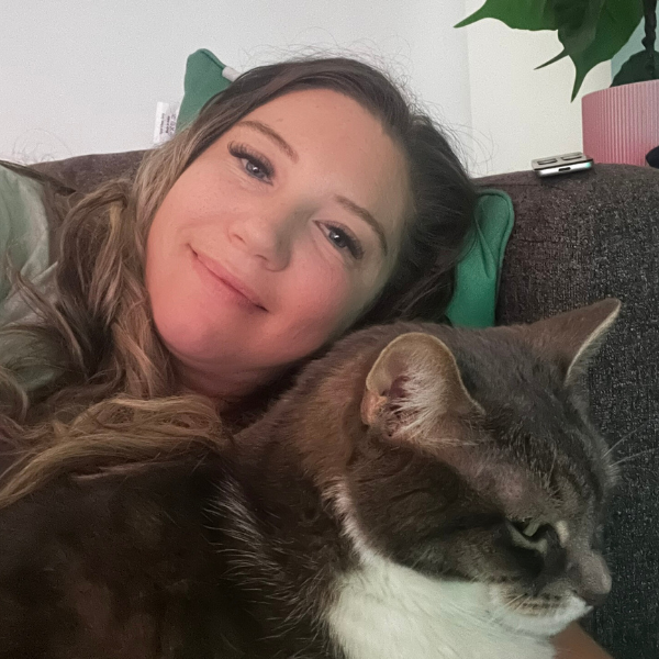 A woman with light skin, long wavy hair, and makeup, smiling while lying on a couch with a gray and white cat.