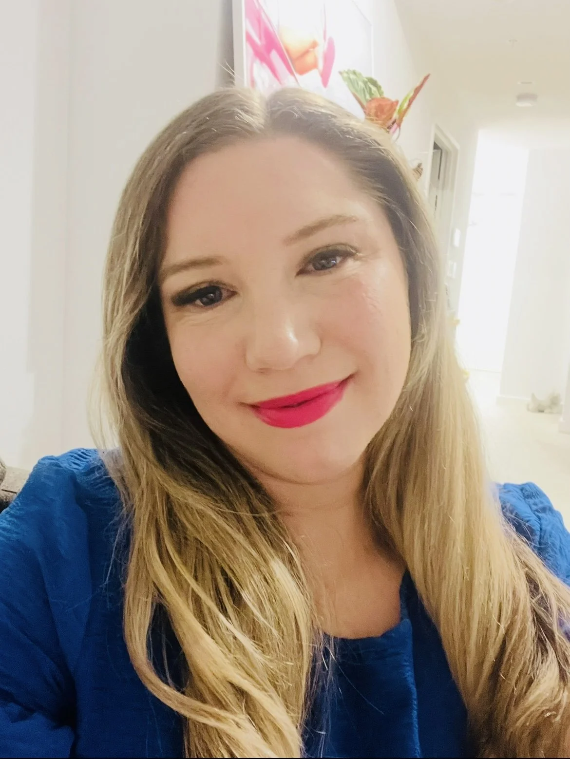 A woman with long blonde hair wearing a blue top, smiling with bright pink lipstick, in a well-lit indoor setting.