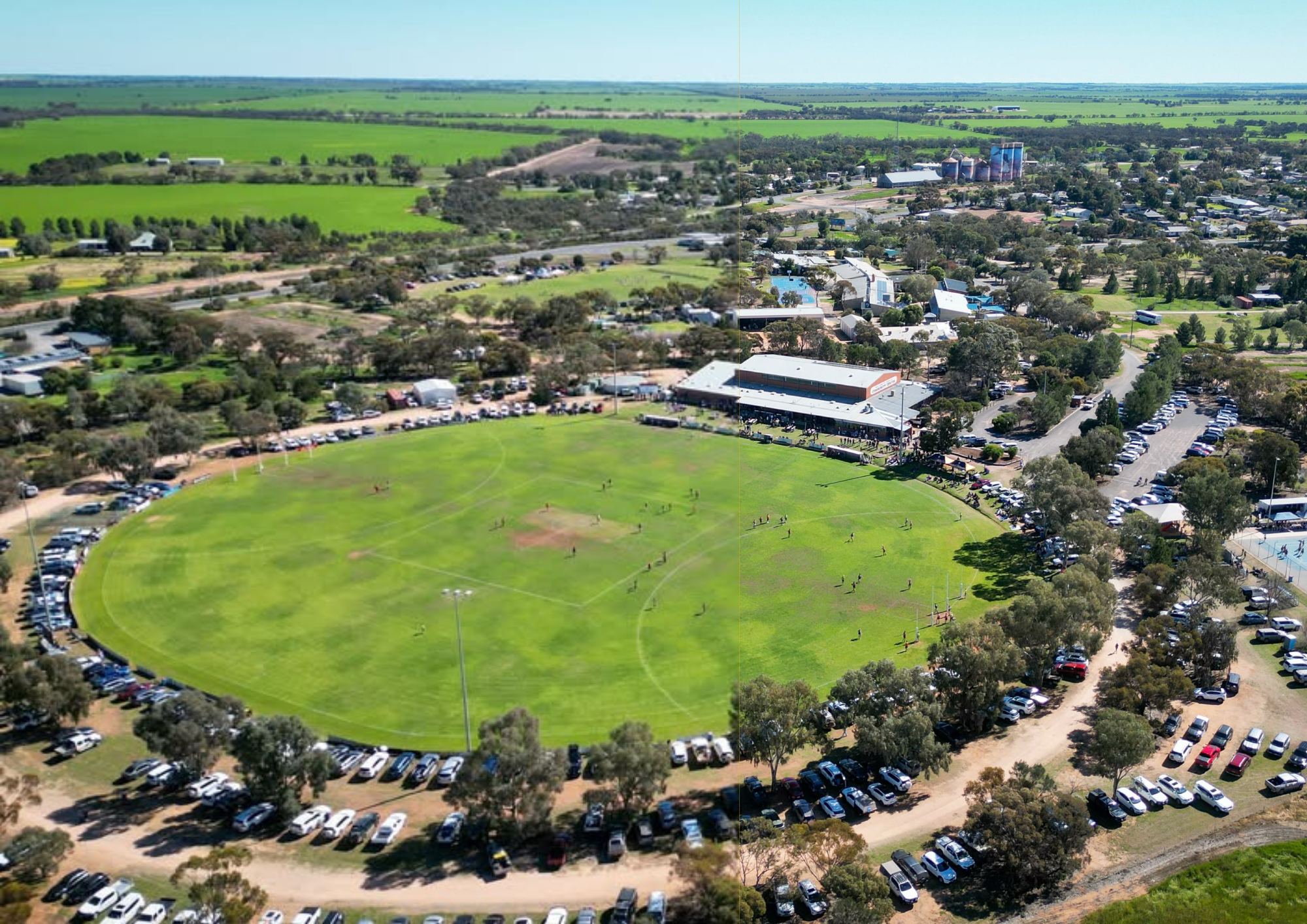 Aerial view of a park with a baseball field and surrounding parking lot, adjacent to a small town with residential and commercial buildings, and green farmland in the background.