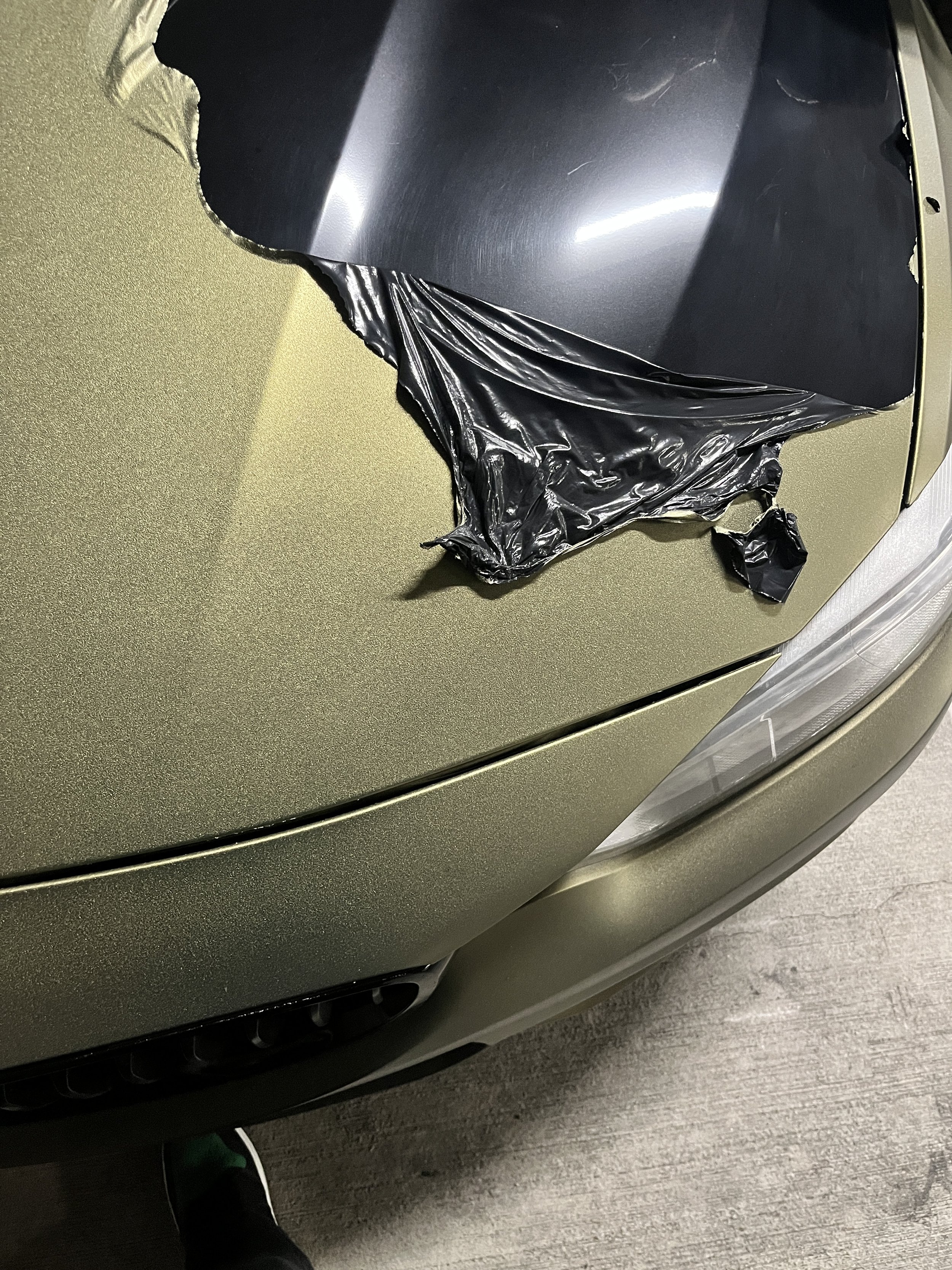 Close-up photo of a green car with a black plastic piece or cover partially peeled back from the hood.