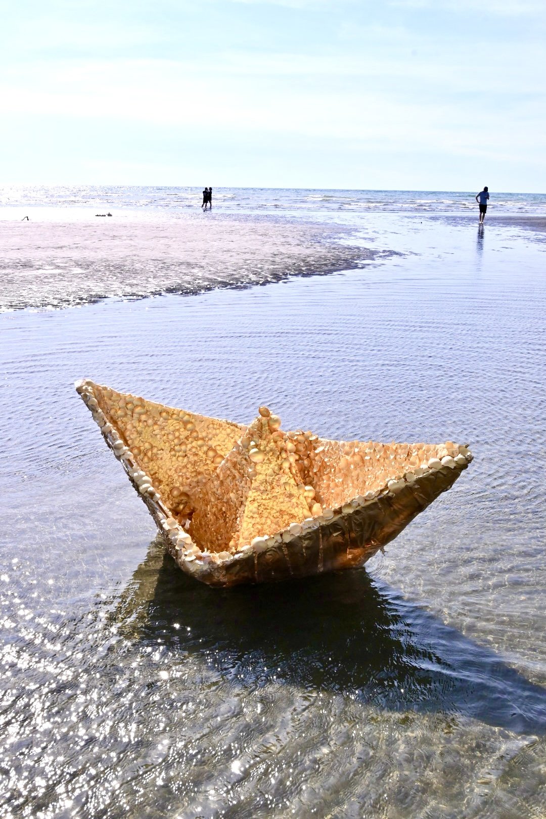 A paper boat made of seashells floating in clear shallow water on a beach with people in the background and the ocean in the distance.
