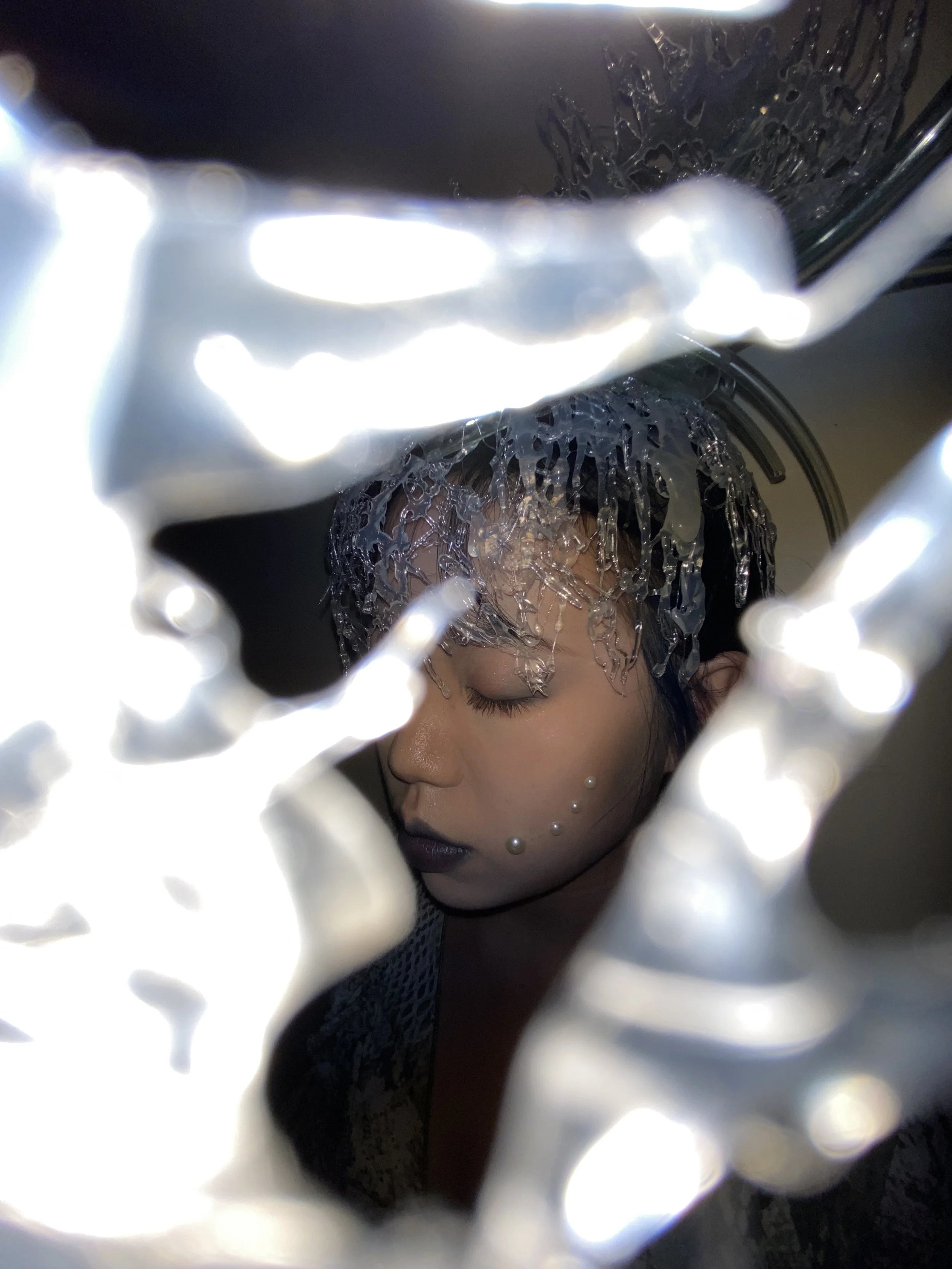 A woman with makeup and pearl face stickers, wearing a translucent, textured headpiece, seen through a reflective, shiny, textured frame.