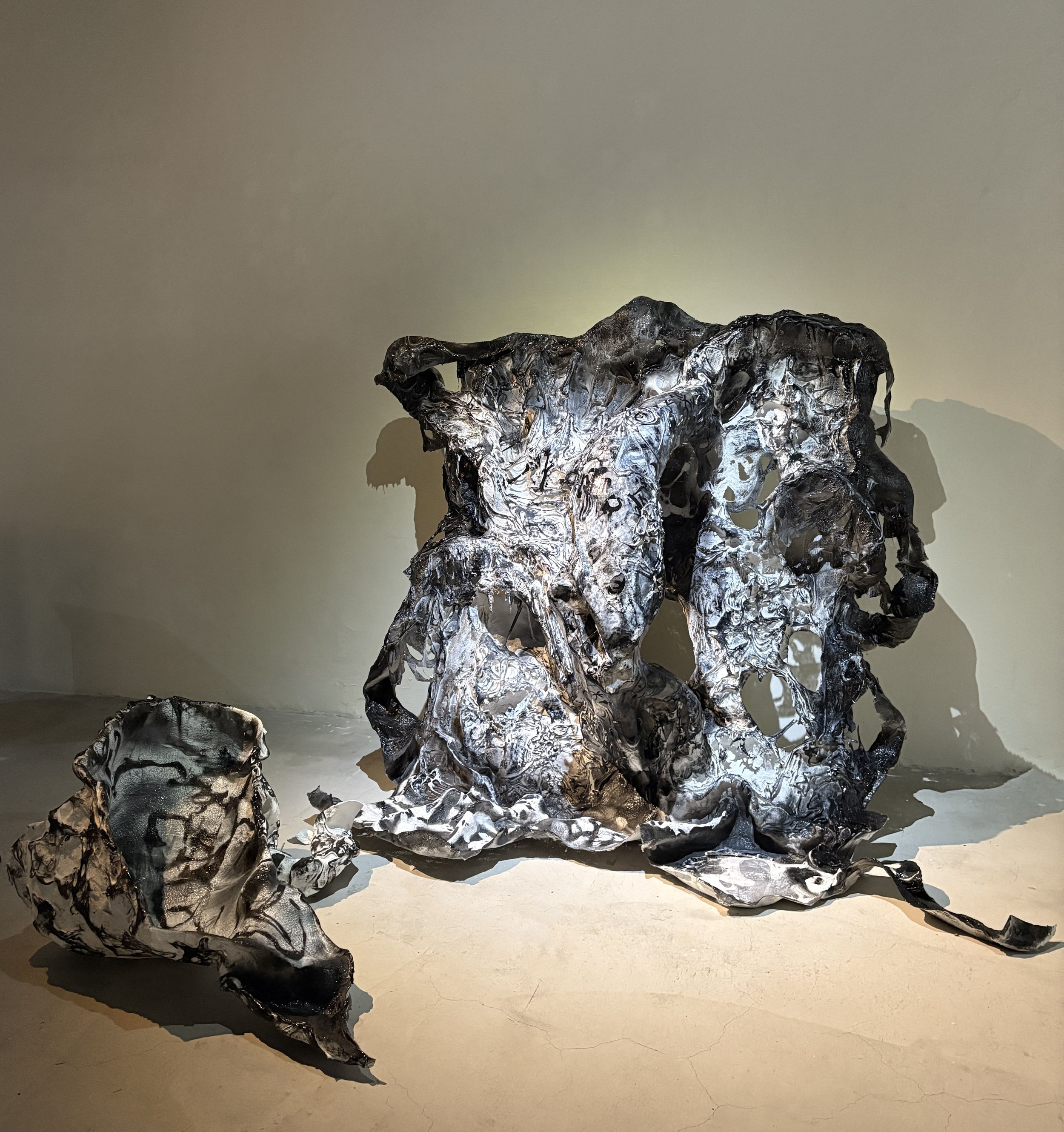 Abstract black and silver sculpture with twisted, organic shapes, placed on a light-colored floor and casting shadows on a beige wall.