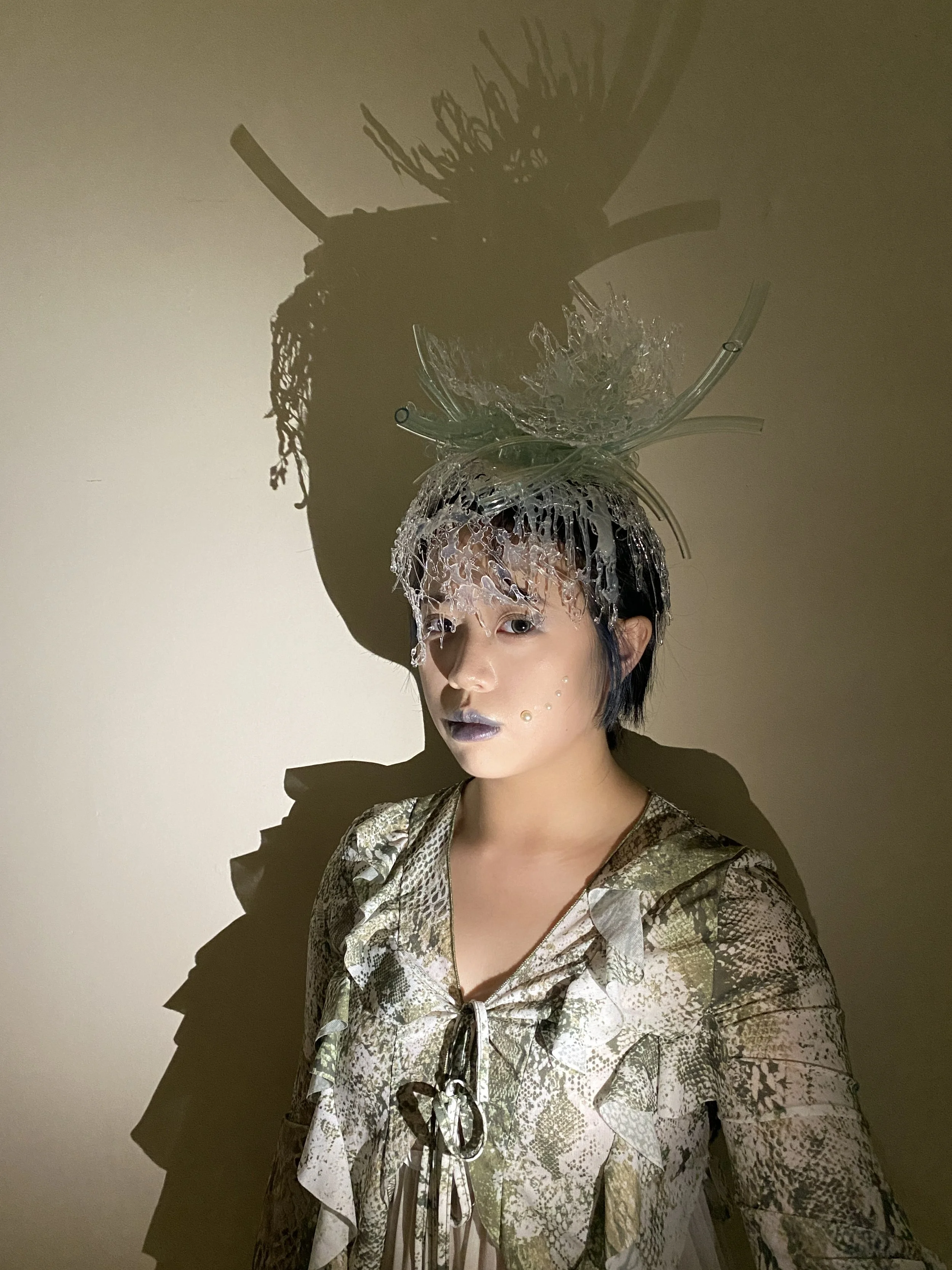 A woman wearing a snake-print dress and an elaborate headpiece made of glass and plastic, with artistic makeup and a shadow cast on a wall behind her.