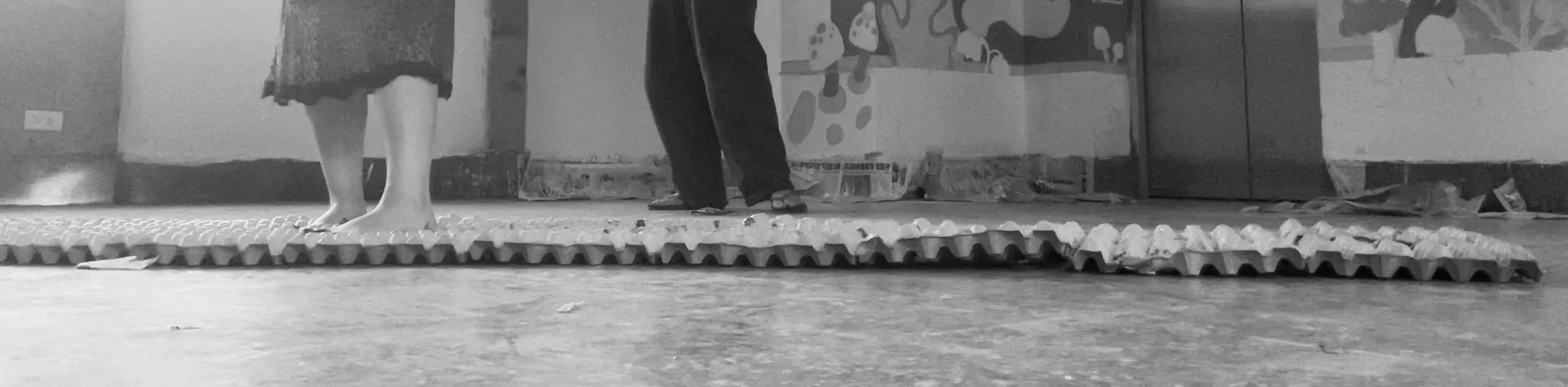 People standing on textured ground mat in an indoor space, with one person barefoot and another wearing sandals, near a painted wall with fungi and tree artwork.
