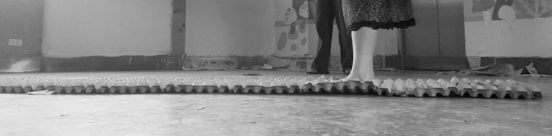 Close-up of two legs of a person standing on a textured foam mat, with a piece of paper and other objects on the floor, and a wall with various images and objects behind them.
