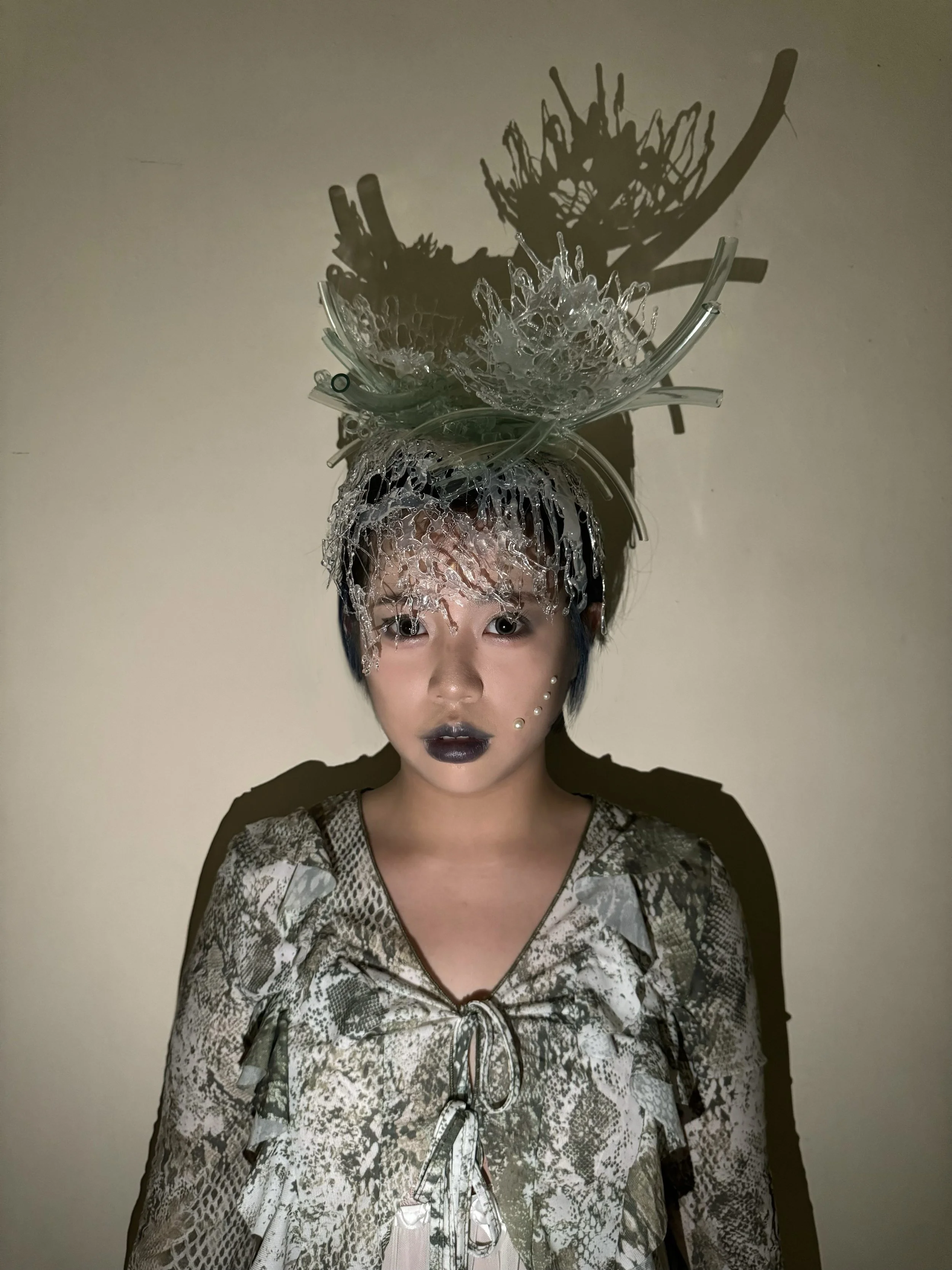 A person with elaborate headwear made of glass or crystal pieces, wearing dark lipstick and a patterned top, standing against a plain wall, with shadows of the headwear cast on the wall.