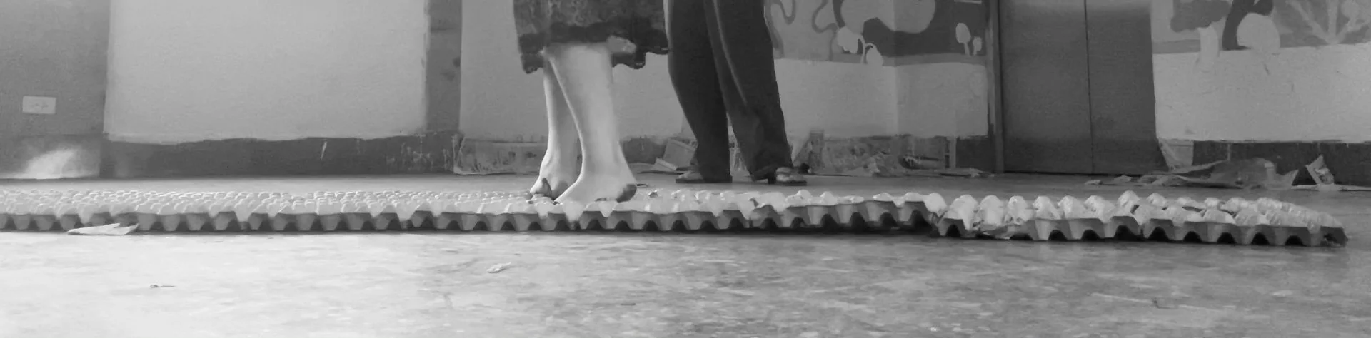 Black and white photo of two people standing on a textured rubber mat, with one person's legs visible from the knees down and the others partially visible behind them.