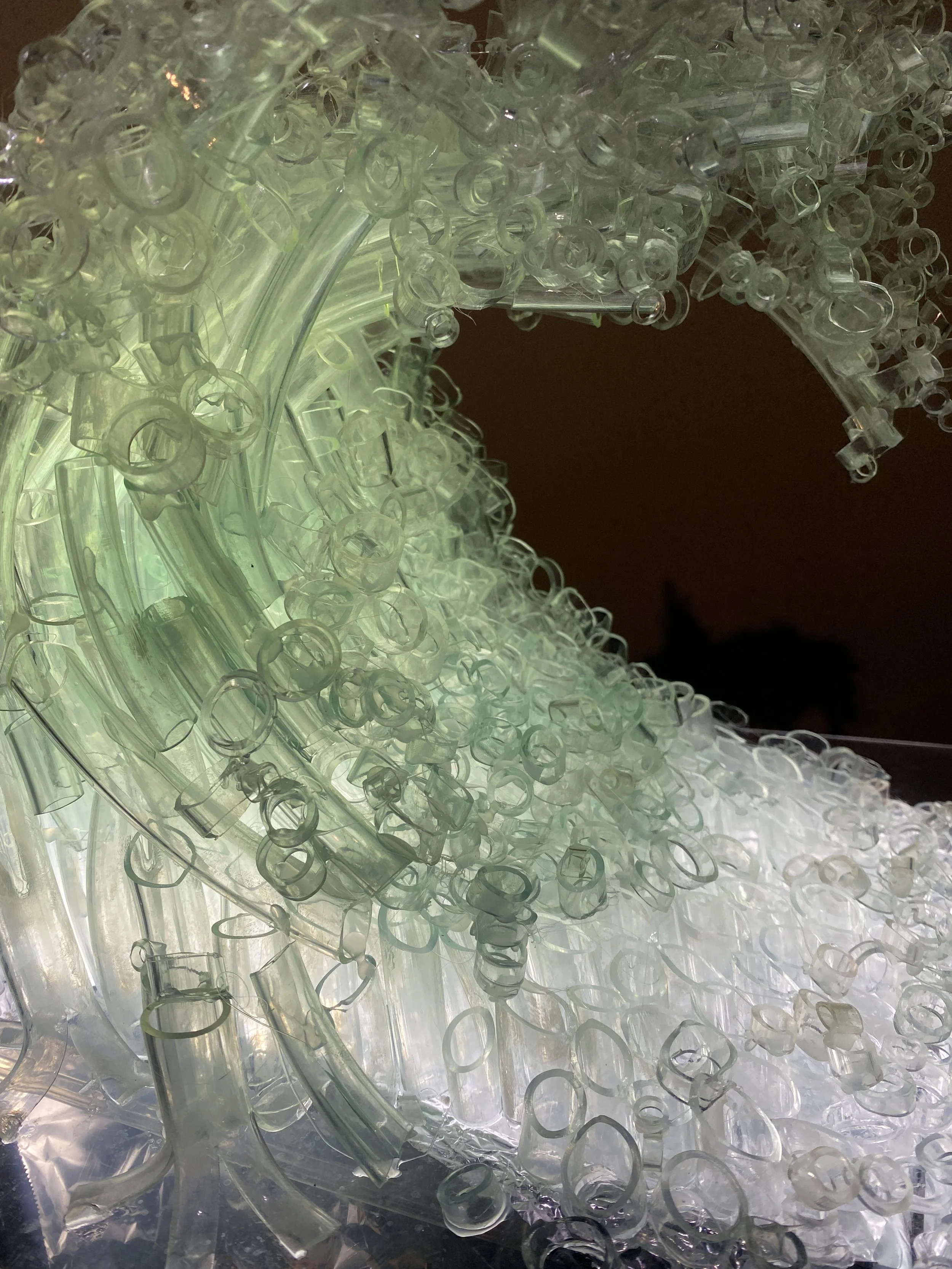 Close-up of a structure made from numerous transparent plastic bottle caps and straws, illuminated from within, with a dark background.