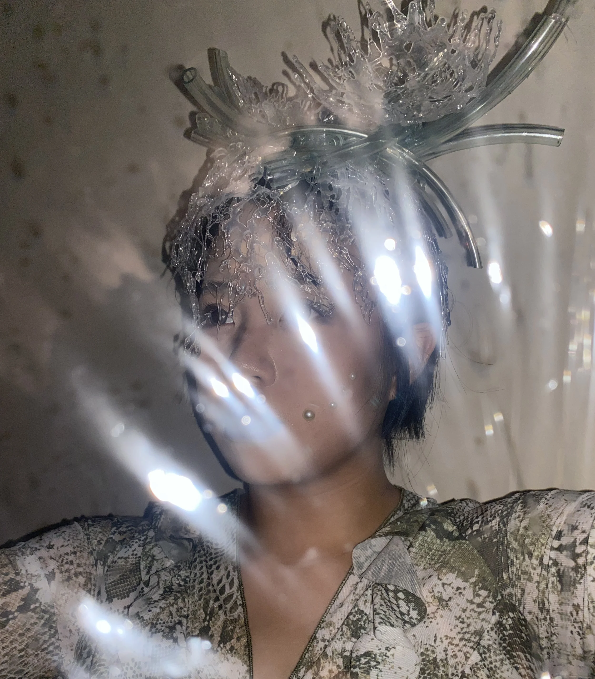 A person with short dark hair wearing an intricate headpiece made of clear plastic and glass-like materials, and a patterned top, standing behind a reflective surface with light reflections creating a shimmering effect.