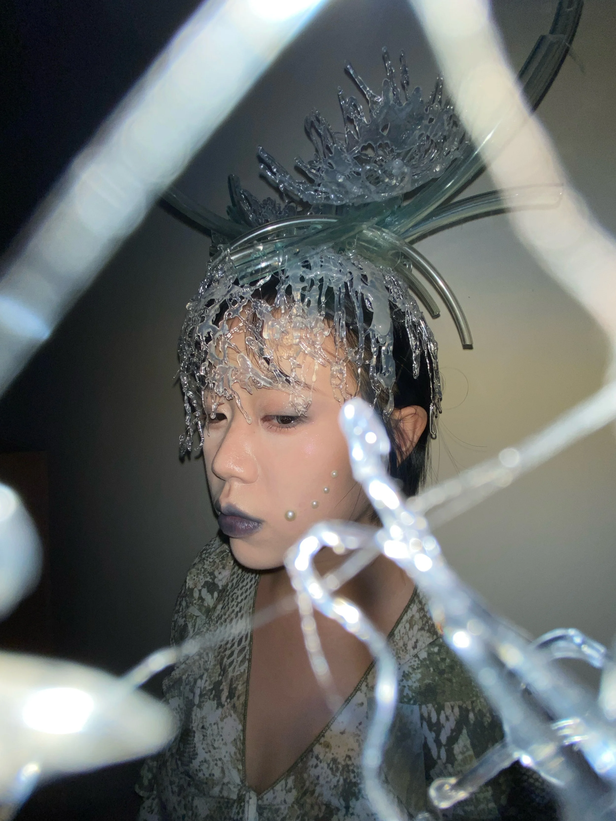 A person with makeup and a metallic headpiece viewed through a glass structure with decorative lights.