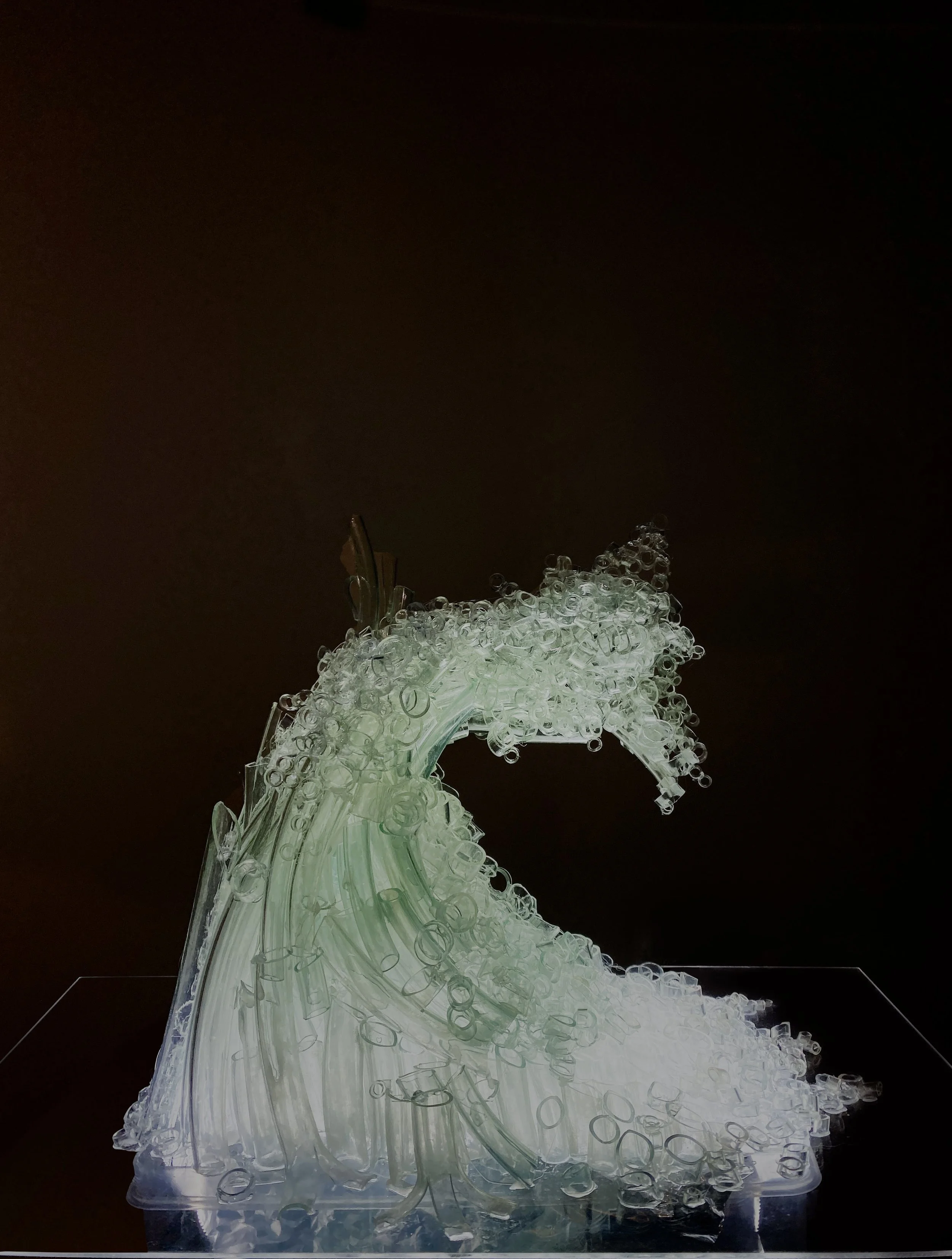 An intricate glass sculpture resembling a wave, made of clear glass tubes and bubbles, mounted on a transparent base against a black background.