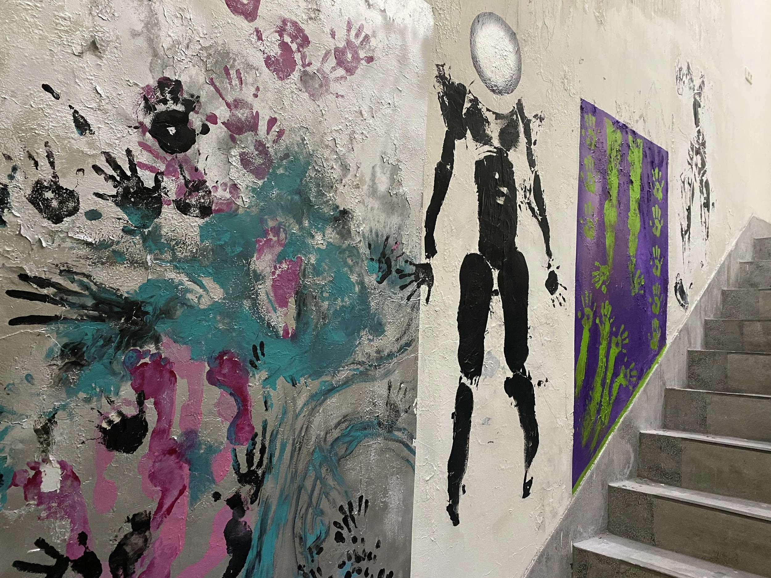 Street art on a wall near stairs, including colorful handprints, a black abstract humanoid figure, and a purple and green artwork with handprints.