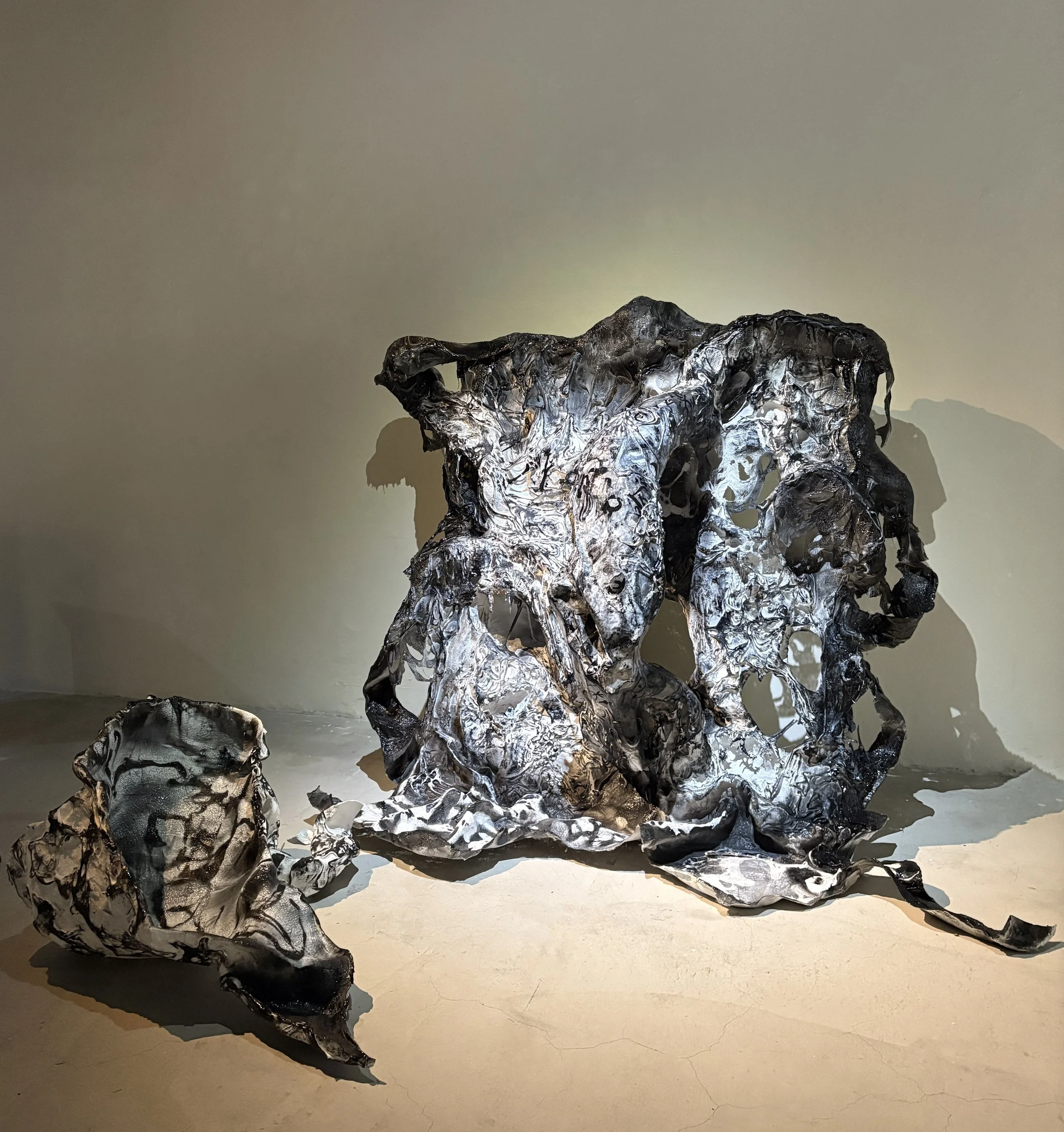 Abstract metal sculpture in black, gray, and silver tones with an irregular, textured surface, displayed against a plain wall with shadows cast on it.