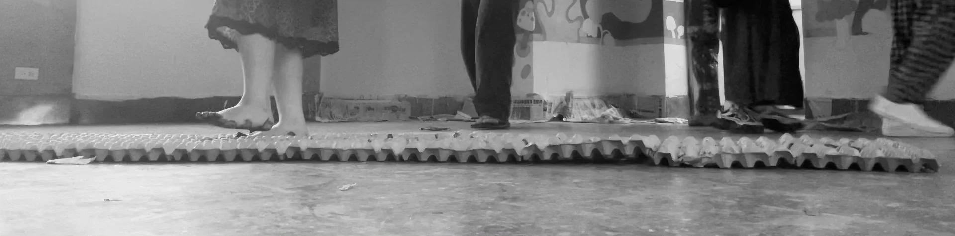 People are walking on a narrow, textured surface in an indoor space, with boxes and artwork visible in the background.