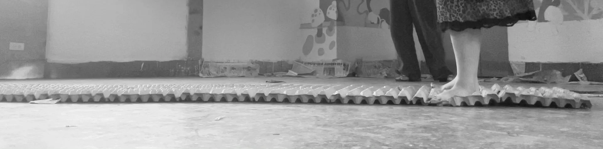 A person standing barefoot on a textured surface, with part of another person visible in the background, in an indoor setting with clutter and artwork on the walls.