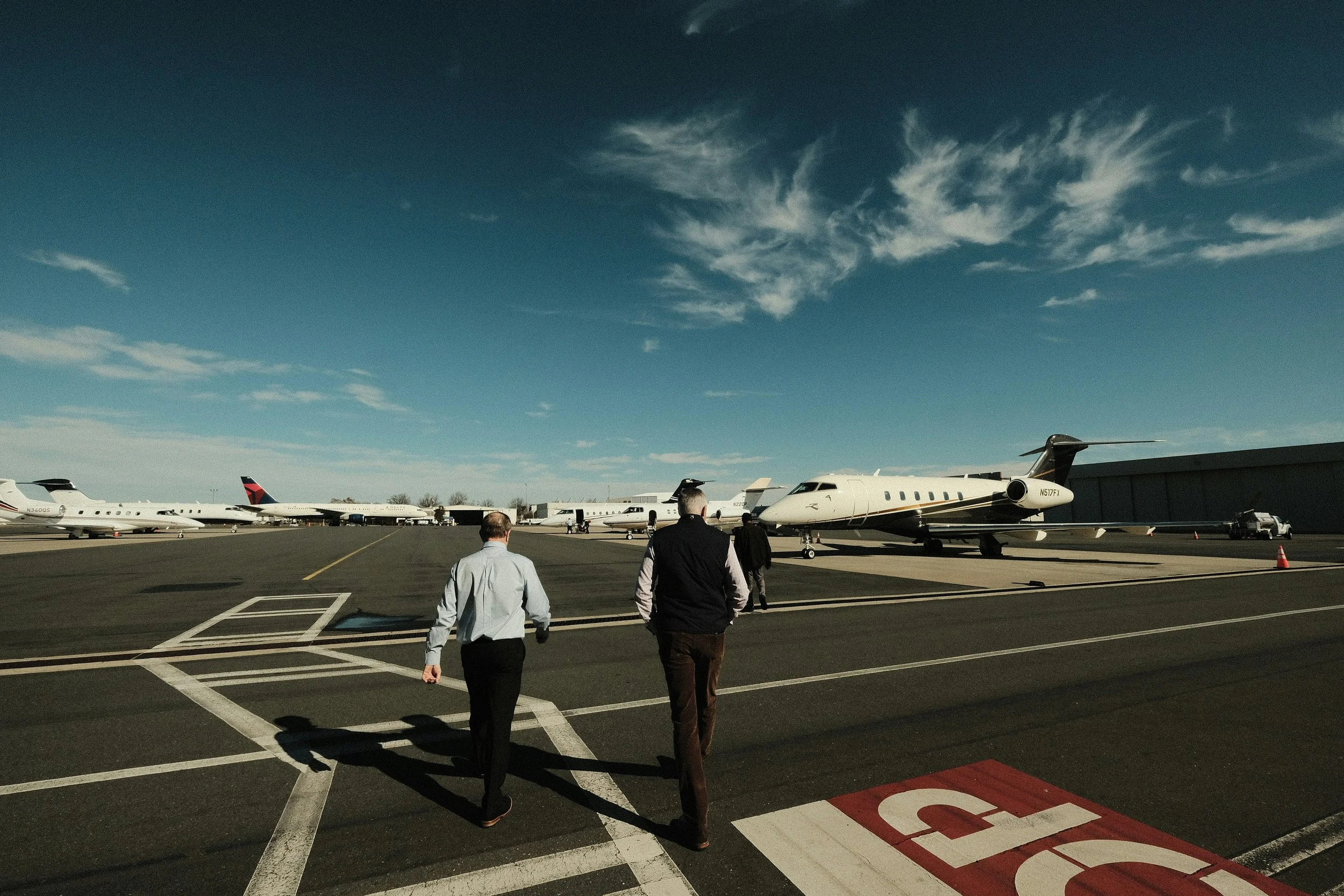 Men walking on tarmac towards private jets parked on an airport runway under a blue sky.