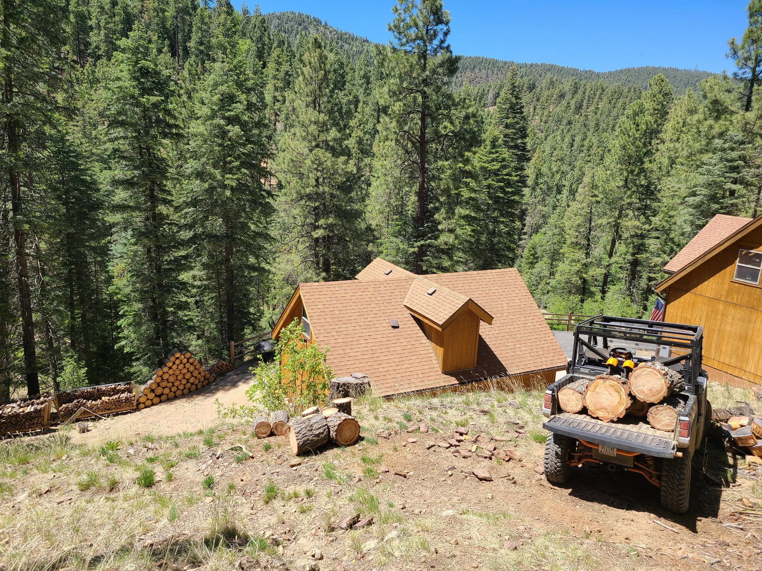 A mountain cabin in a forested area with trees in the background. There is a flat area in front with logs stacked on the side, and a utility vehicle carrying large logs parked on the right.