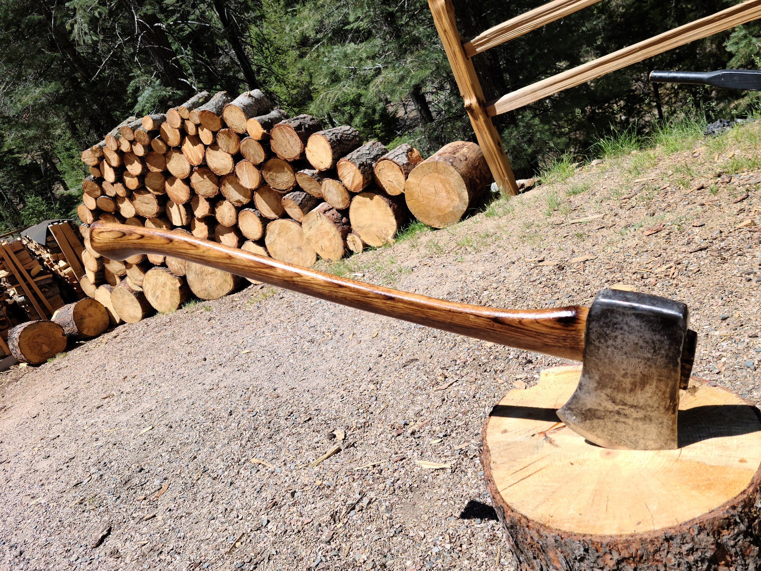 An axe embedded in a wooden block in a gravel area next to a stack of chopped firewood and a wooden chart