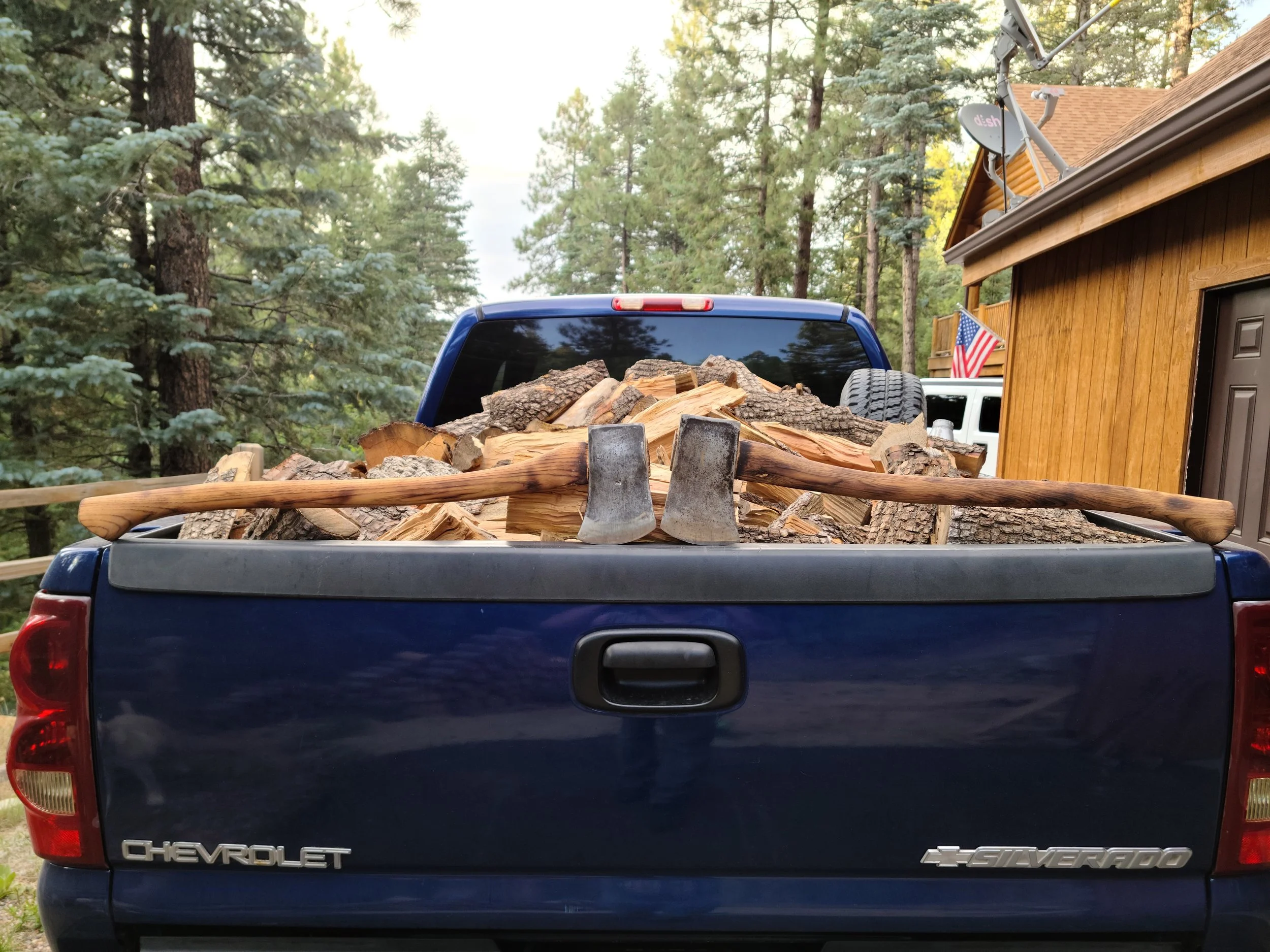 A blue Chevrolet Silverado pickup truck loaded with chopped firewood in the truck bed, parked outdoors near trees and a wooden building.