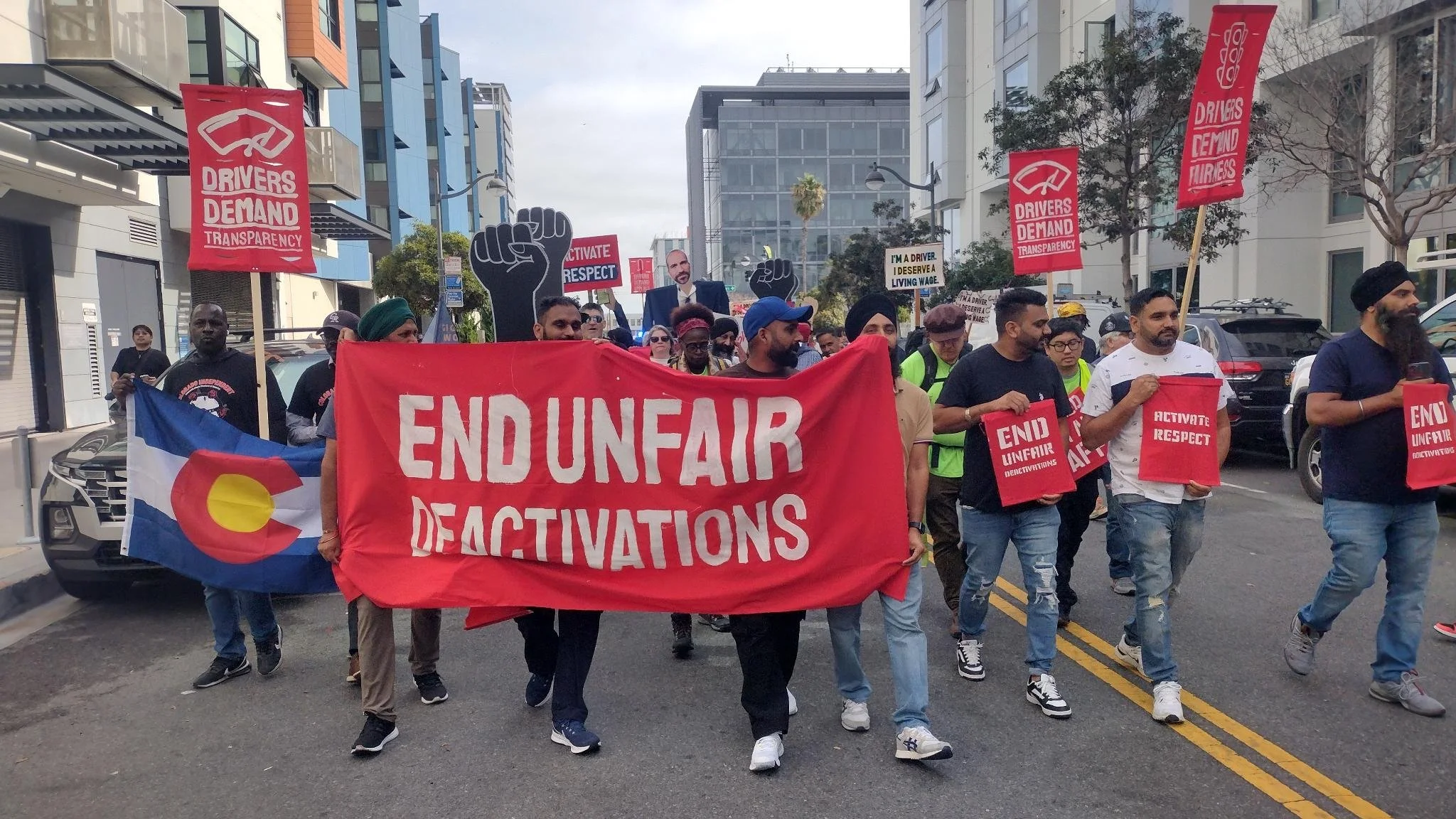 A group of protesters marching down a city street holding signs and banners advocating for driver rights and ending unfair deactivations. Some signs read 'Drivers Demand Transparency', 'End Unfair Deactivations', and 'Activate Respect'. One protester is holding an Australian flag with a red and yellow symbol.