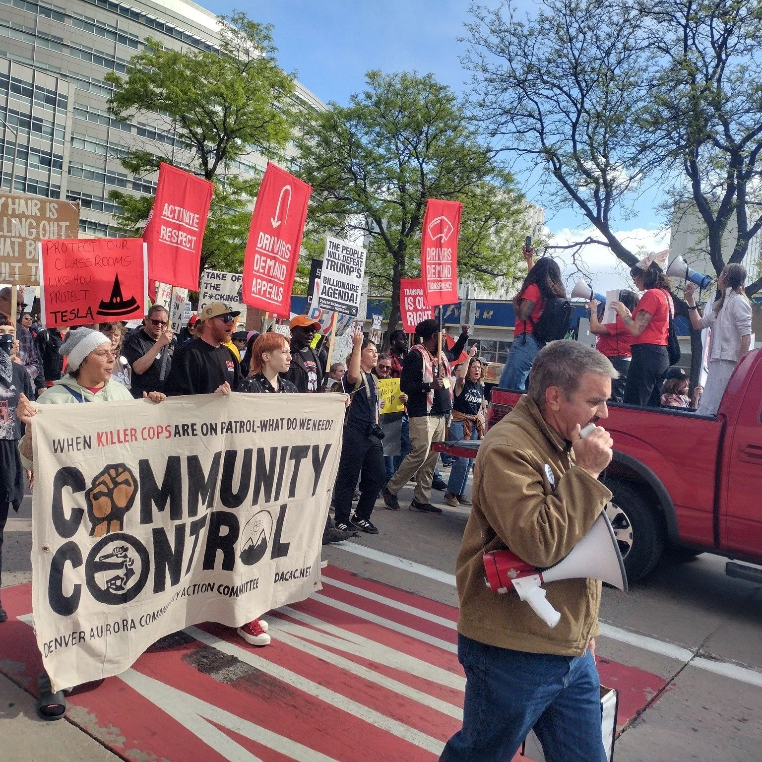 A protest march with people holding various signs and banners. One prominent banner reads "Community Control" with icons and below it, "When killer cops are on patrol - what do we need?" The marchers are walking on a crosswalk and some are speaking into megaphones. People are gathered near a building with trees around.