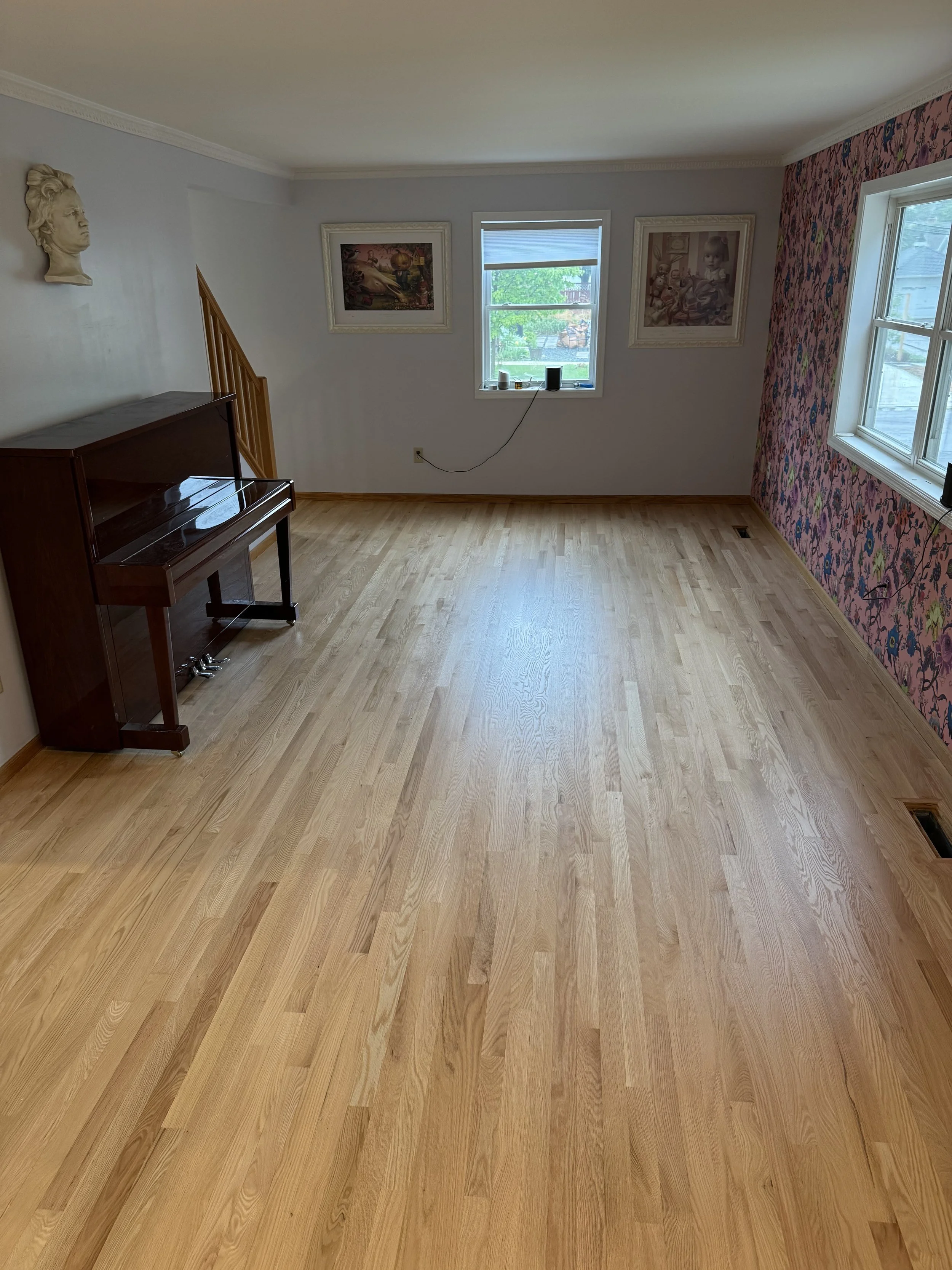 Empty living room with light wood flooring, painted walls, two framed artworks, a pink and black patterned wall, and a small upright piano in the corner.