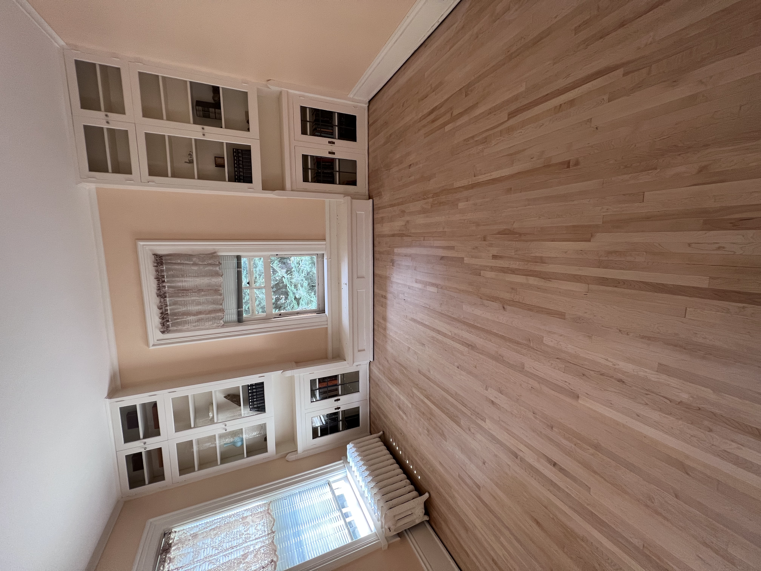 Empty room with wooden floors, white built-in bookshelves, a window with curtains, and a radiator.