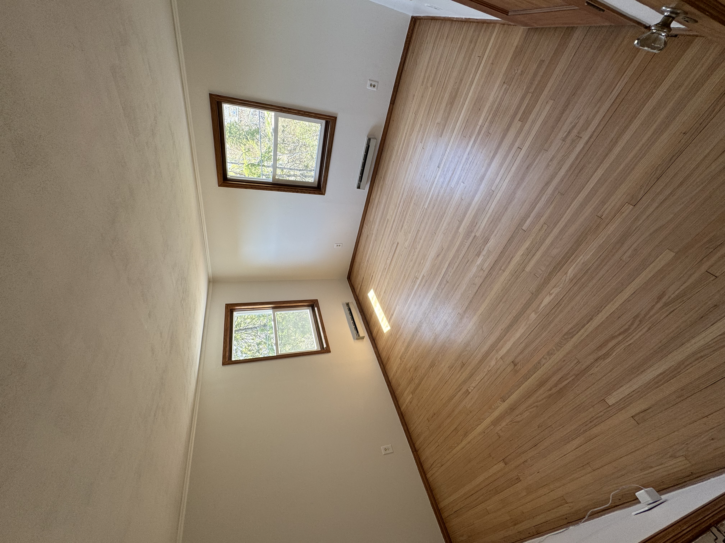 Empty room with hardwood floors, white walls, two windows with wooden frames, and air conditioning units under each window.