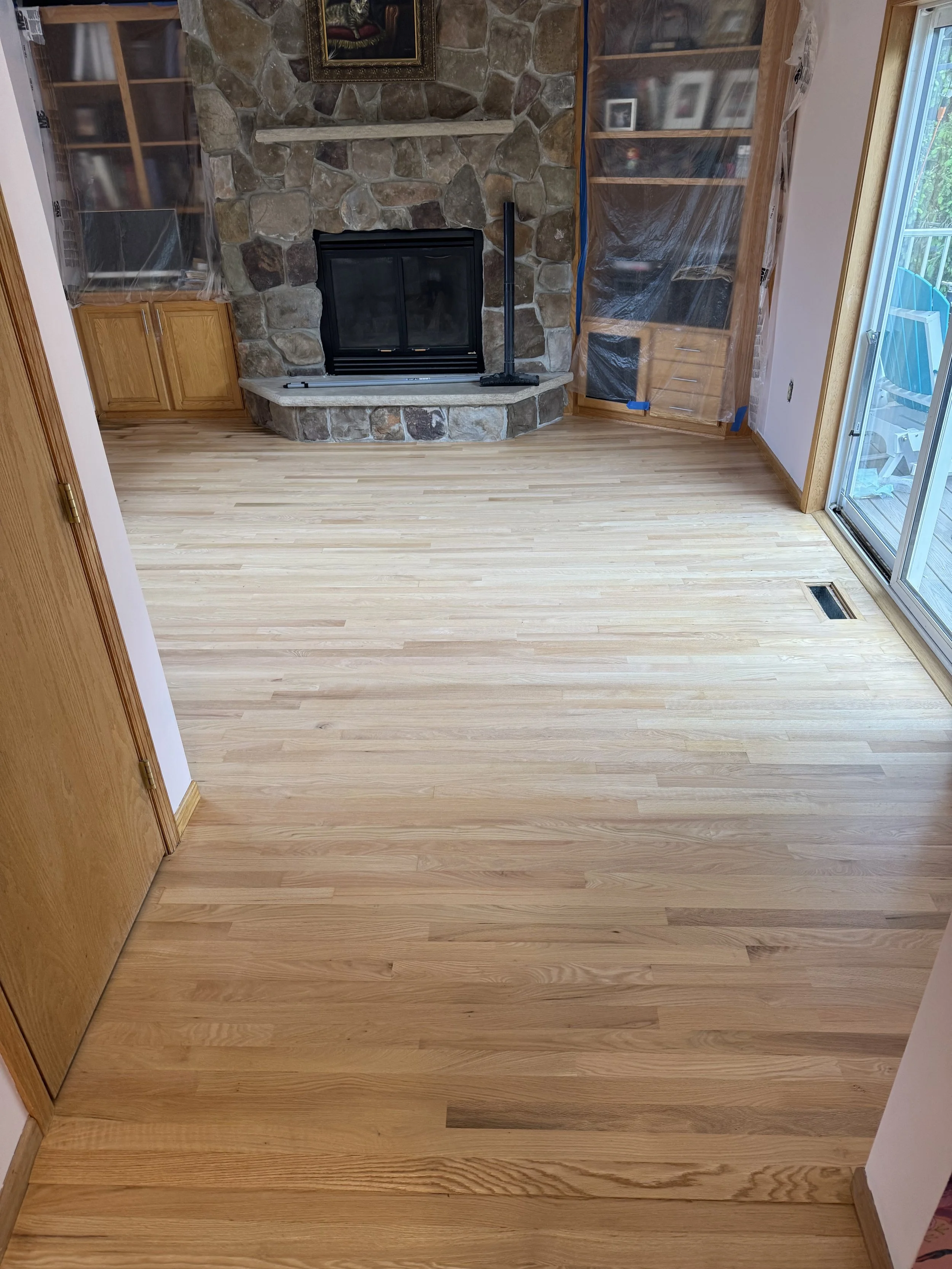 Empty living room with light wood flooring, stone fireplace, built-in cabinets, and sliding glass door leading outside.